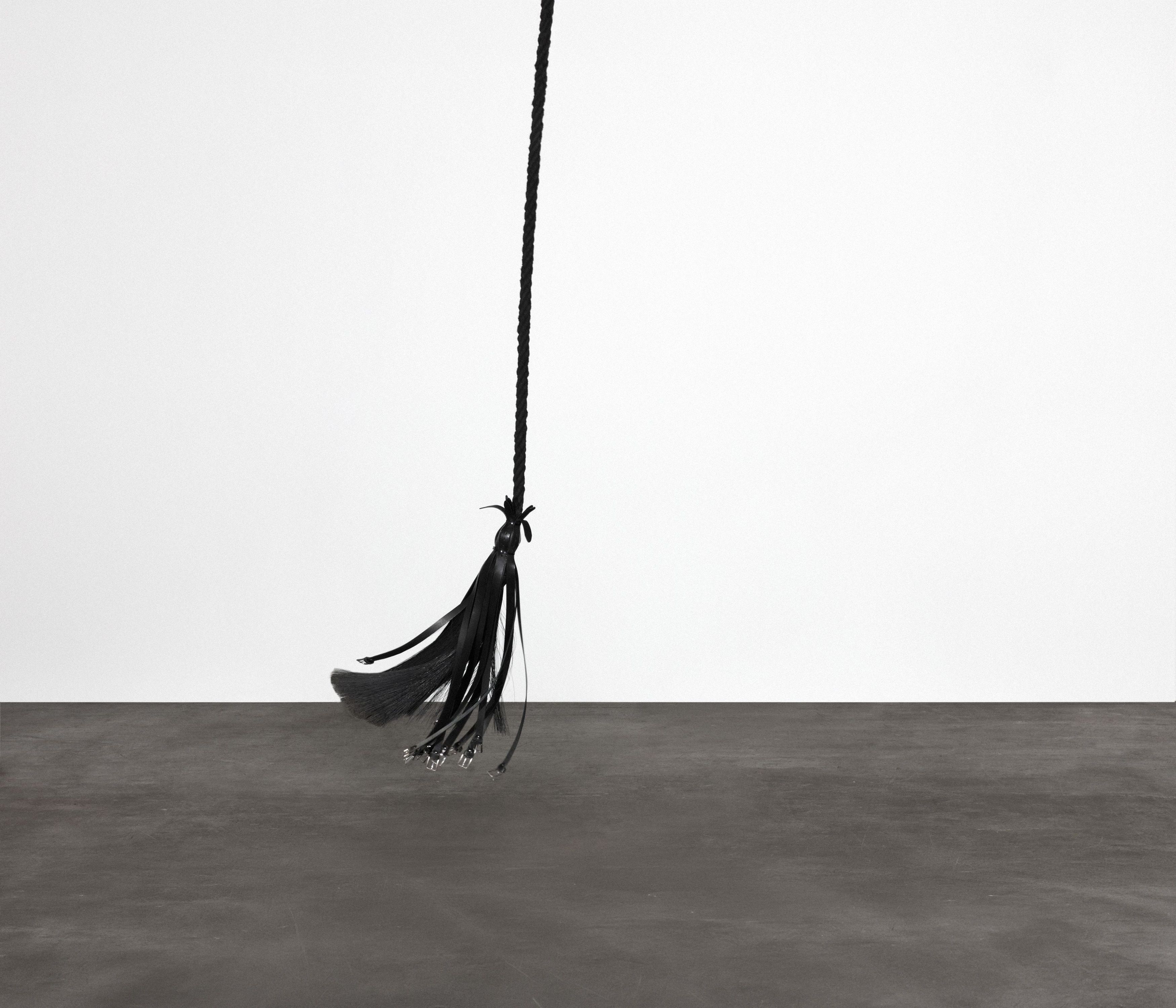 A long, black braided sculpture with a tassel end hangs vertically against a stark white wall above a grey floor.