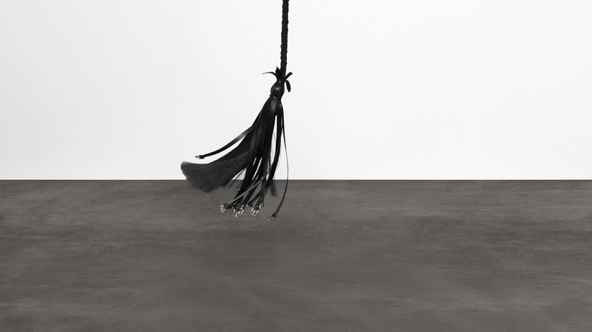 A long, black braided sculpture with a tassel end hangs vertically against a stark white wall above a grey floor.