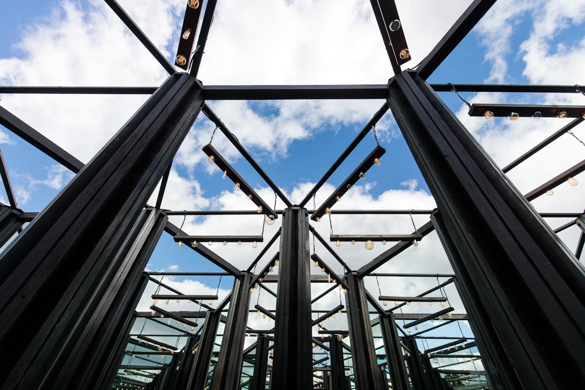 A low-angle shot looking up through a complex, geometric black metal frame fitted with hanging Edison-style light bulbs. The structure creates a mirrored, kaleidoscopic effect against a bright blue sky filled with soft white clouds.