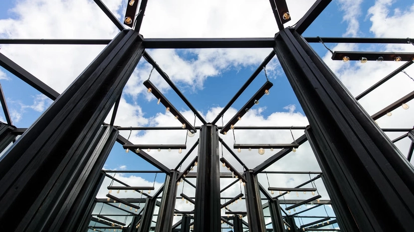 A low-angle shot looking up through a complex, geometric black metal frame fitted with hanging Edison-style light bulbs. The structure creates a mirrored, kaleidoscopic effect against a bright blue sky filled with soft white clouds.