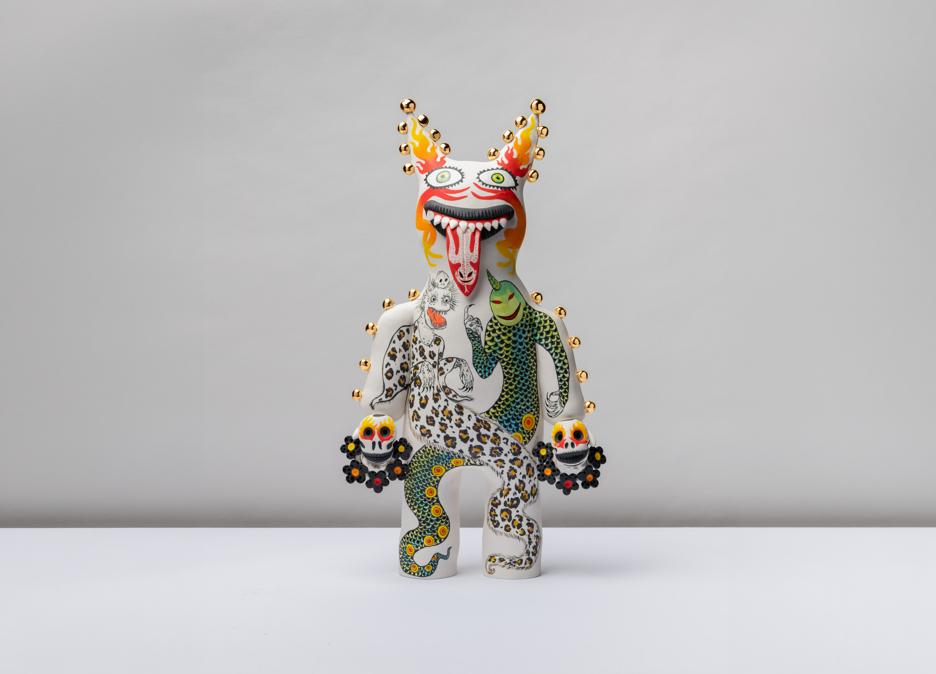 A whimsical, white ceramic figure features a wide, grinning mouth with a long tongue and decorative gold studs lining its body. The sculpture is intricately painted with colorful motifs, including a leopard-print creature and a green scaled figure, while holding small skull-like objects in each hand.
