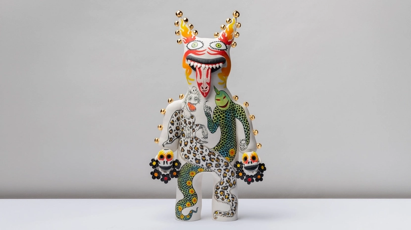 A whimsical, white ceramic figure features a wide, grinning mouth with a long tongue and decorative gold studs lining its body. The sculpture is intricately painted with colorful motifs, including a leopard-print creature and a green scaled figure, while holding small skull-like objects in each hand.