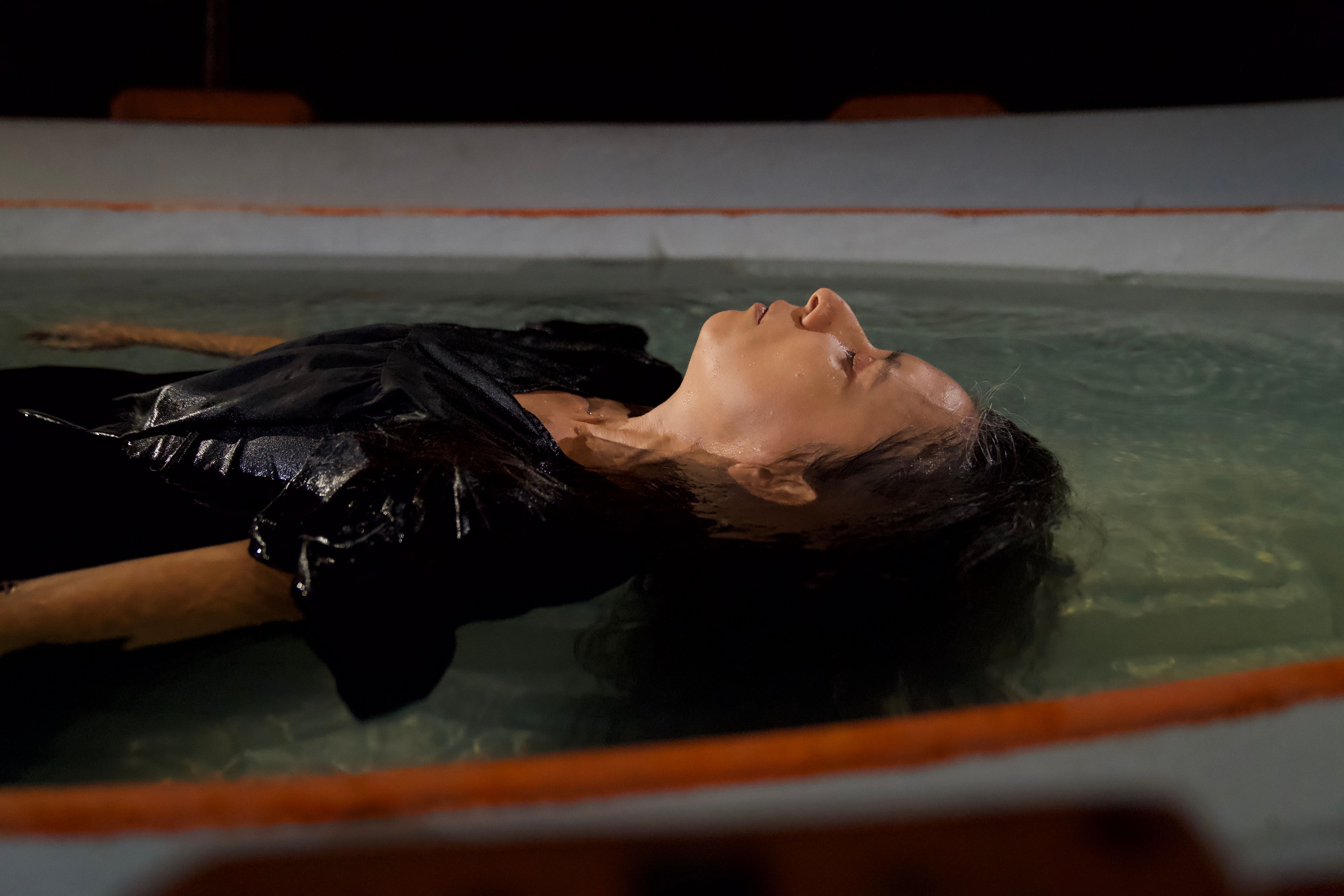 A person in black clothes floats peacefully on their back in a shallow pool of water. The close-up side profile captures their face breaking the surface in a calm, dark environment.