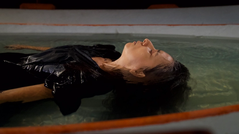 A person in black clothes floats peacefully on their back in a shallow pool of water. The close-up side profile captures their face breaking the surface in a calm, dark environment.