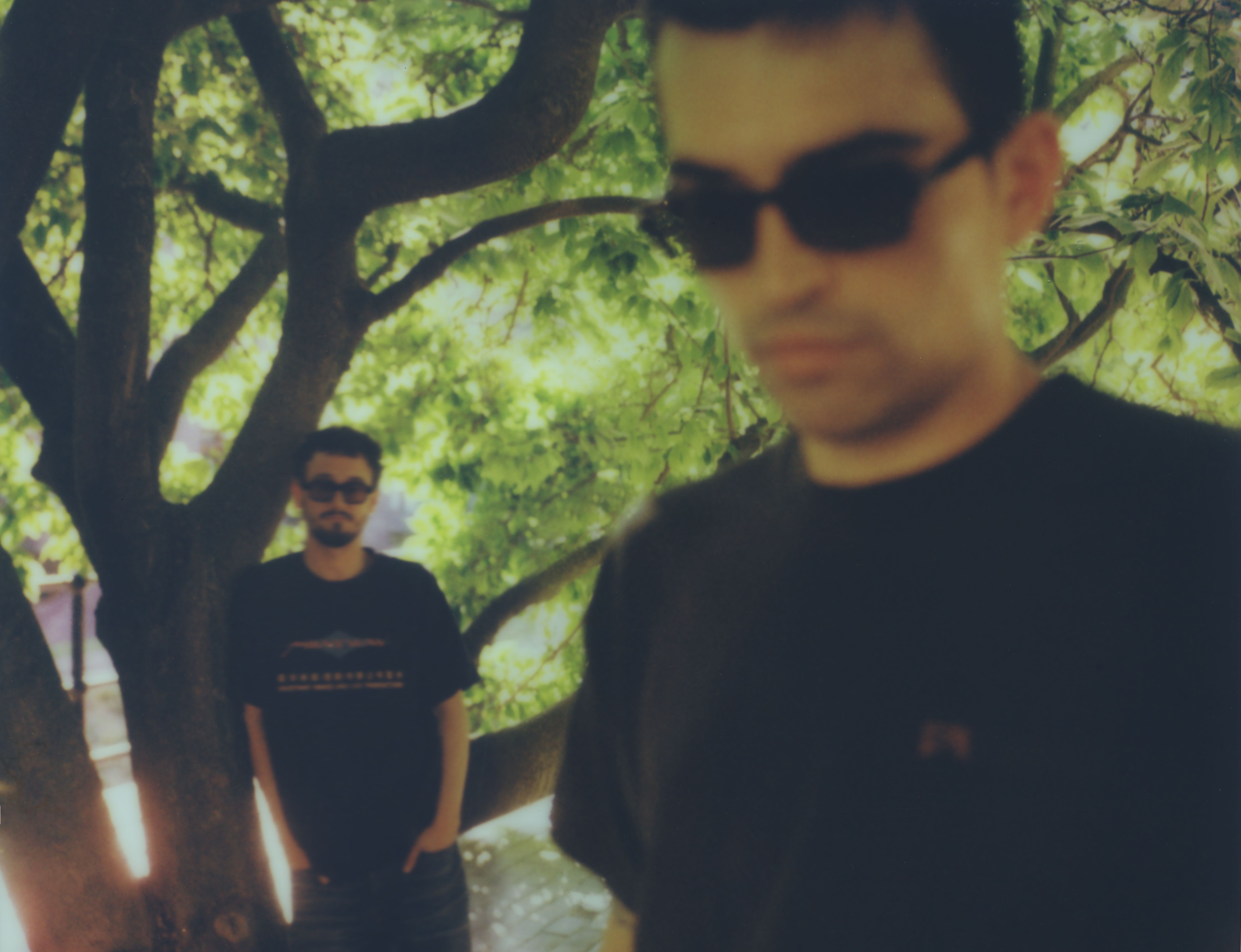 A blurry, low-fidelity photograph of two men wearing sunglasses outdoors near a large tree. One man stands in the background by the tree trunk, while the other is positioned prominently but out of focus in the foreground.