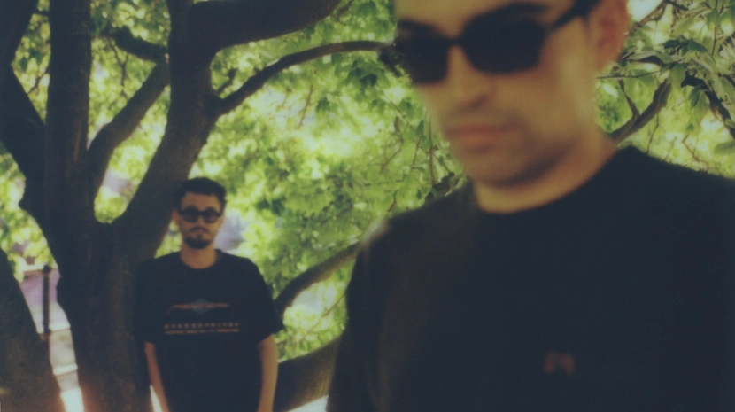 A blurry, low-fidelity photograph of two men wearing sunglasses outdoors near a large tree. One man stands in the background by the tree trunk, while the other is positioned prominently but out of focus in the foreground.