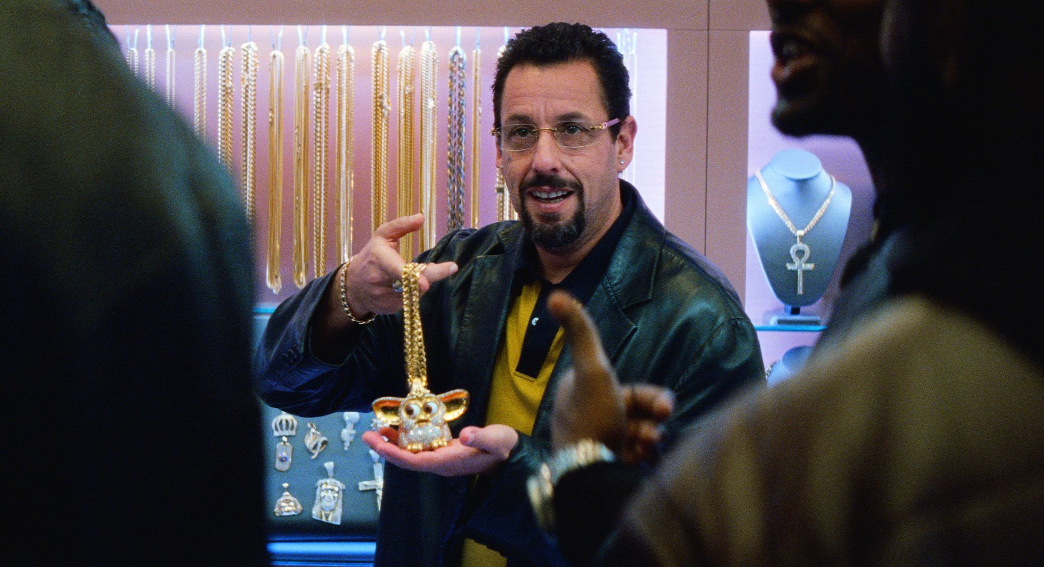 Adam Sandler, wearing tinted glasses and a leather jacket, holds up a large, diamond-encrusted "Furby" pendant in a brightly lit jewelry store. He is surrounded by display cases filled with gold chains and religious charms as he speaks to customers in the foreground.