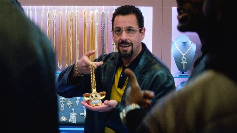 Adam Sandler, wearing tinted glasses and a leather jacket, holds up a large, diamond-encrusted "Furby" pendant in a brightly lit jewelry store. He is surrounded by display cases filled with gold chains and religious charms as he speaks to customers in the foreground.