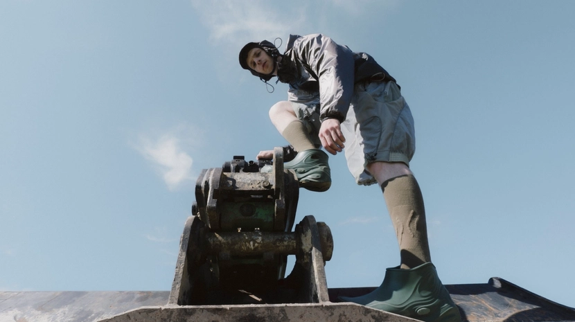 A person wearing a metallic jacket, shorts, and green sculpted boots stands atop a piece of heavy machinery against a clear blue sky. The low-angle shot emphasises their futuristic, utilitarian fashion and the industrial equipment.