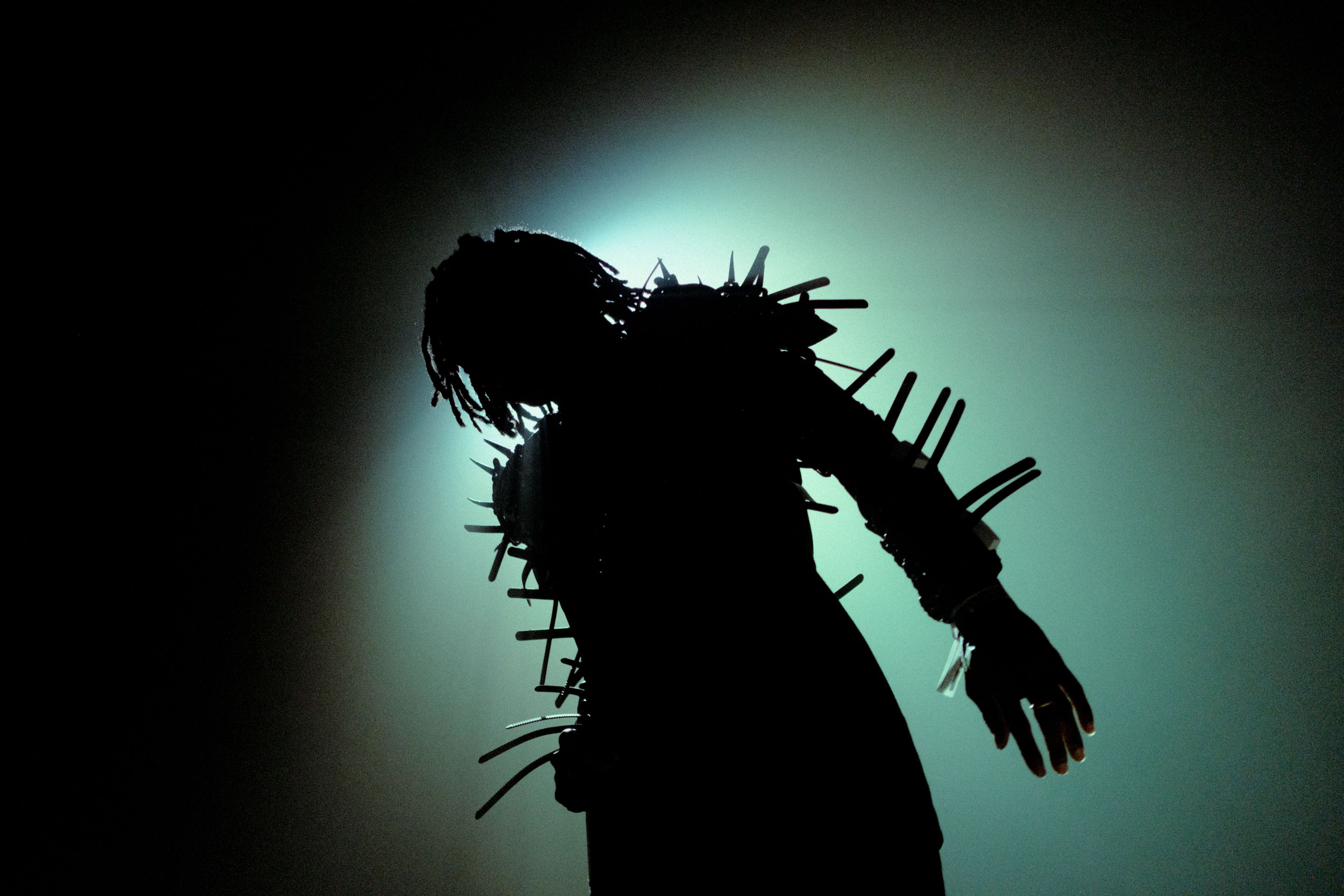 A dark silhouette of a person wearing a garment covered in long, protruding spikes stands against a hazy green backlight. The dramatic lighting hides their features while highlighting the aggressive, textured shape of their attire.