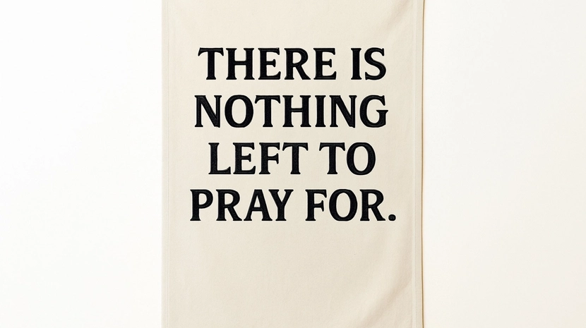 A minimalist textile art piece featuring a cream-colored fabric banner with the words "THERE IS NOTHING LEFT TO PRAY FOR." embroidered in bold, black capital letters. The banner hangs flat against a plain, off-white wall.