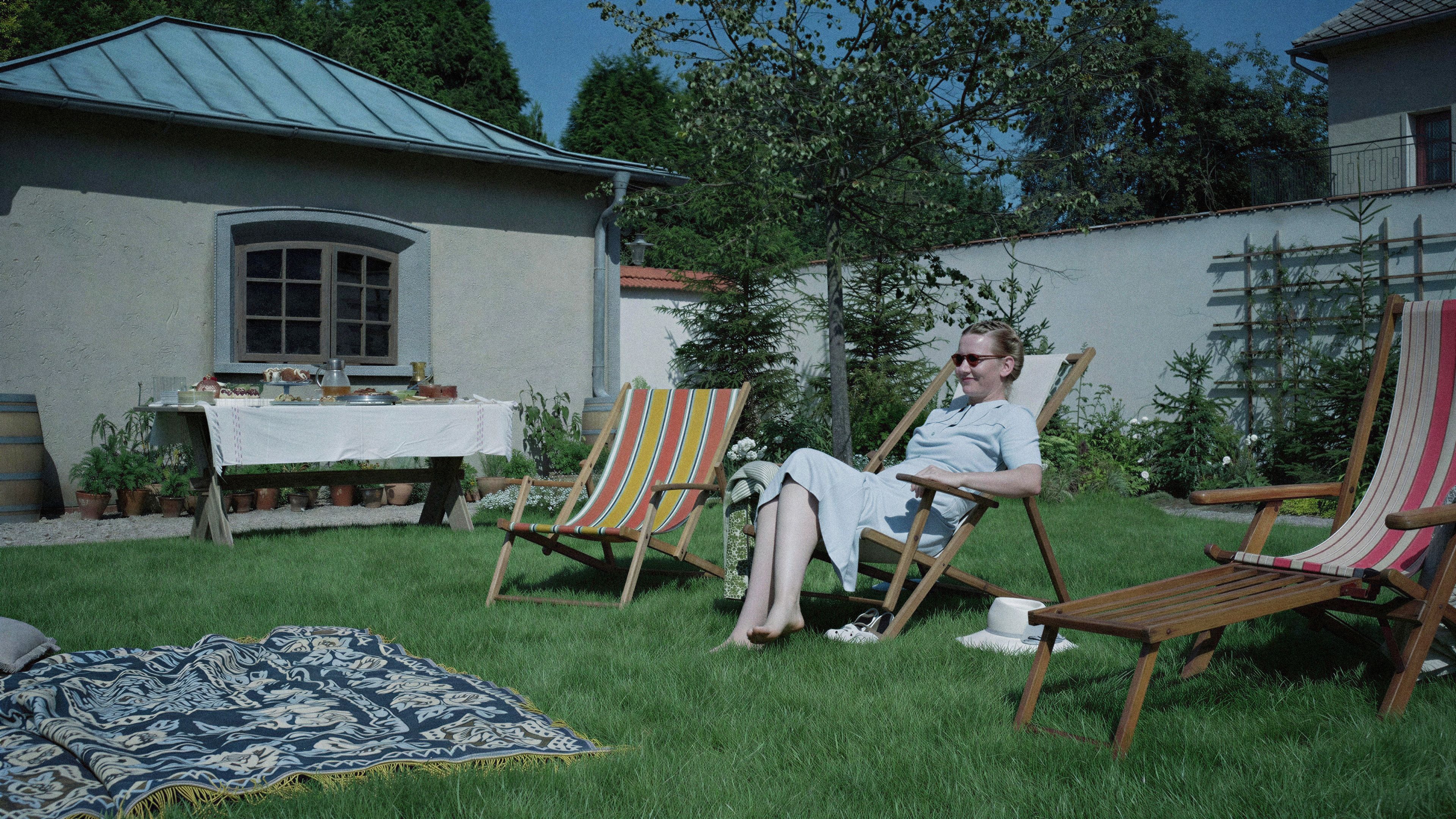 A woman in a blue dress and sunglasses relaxes in a wooden lounge chair on a sunny lawn. In the background, a white-clothed table is laden with food next to a small stone building.