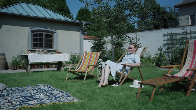 A woman in a blue dress and sunglasses relaxes in a wooden lounge chair on a sunny lawn. In the background, a white-clothed table is laden with food next to a small stone building.
