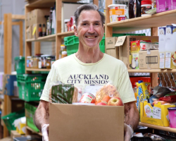 a man in an auckland city mission shirt holds a box of food