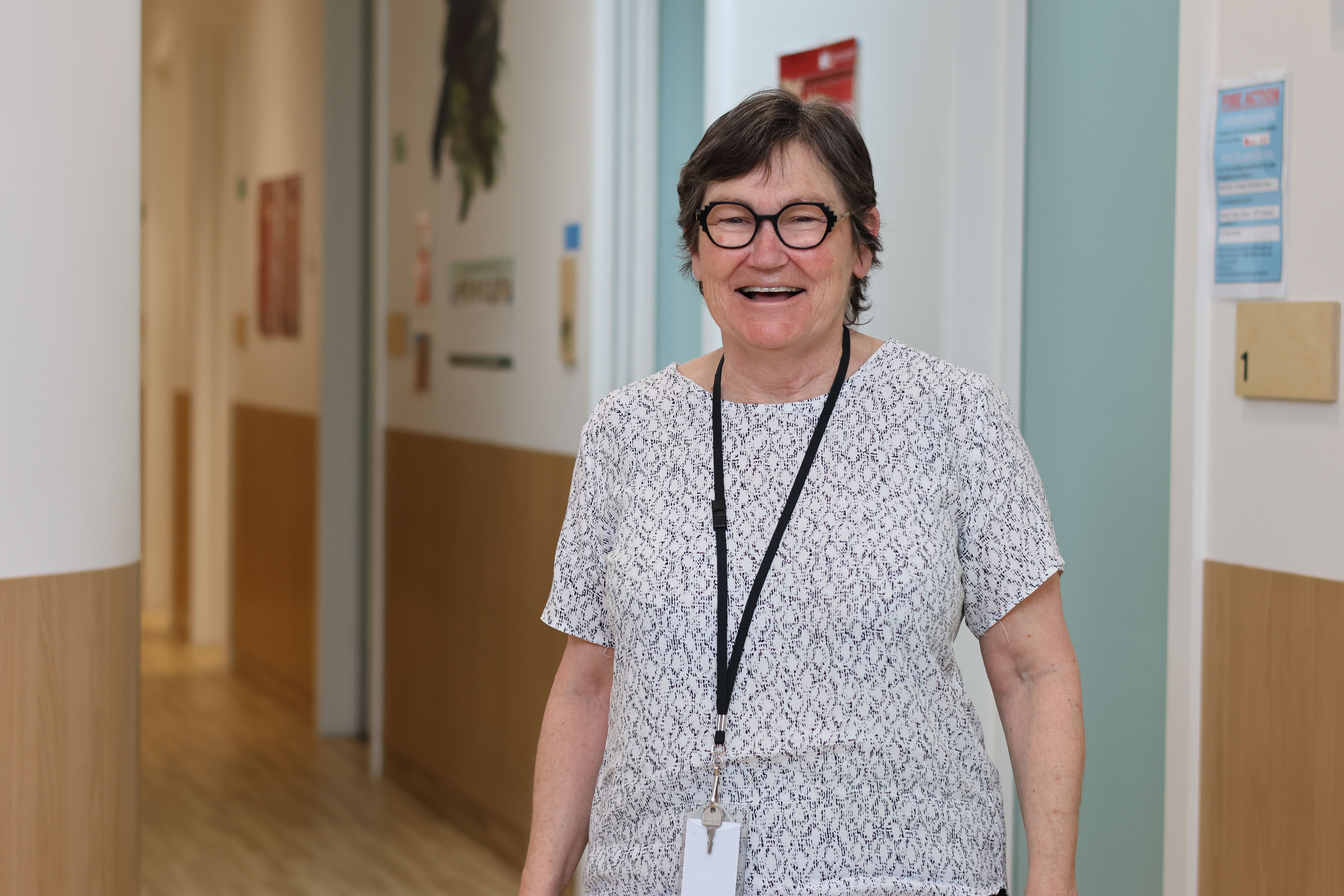 a woman wearing glasses and a lanyard is standing in a hallway and smiling .