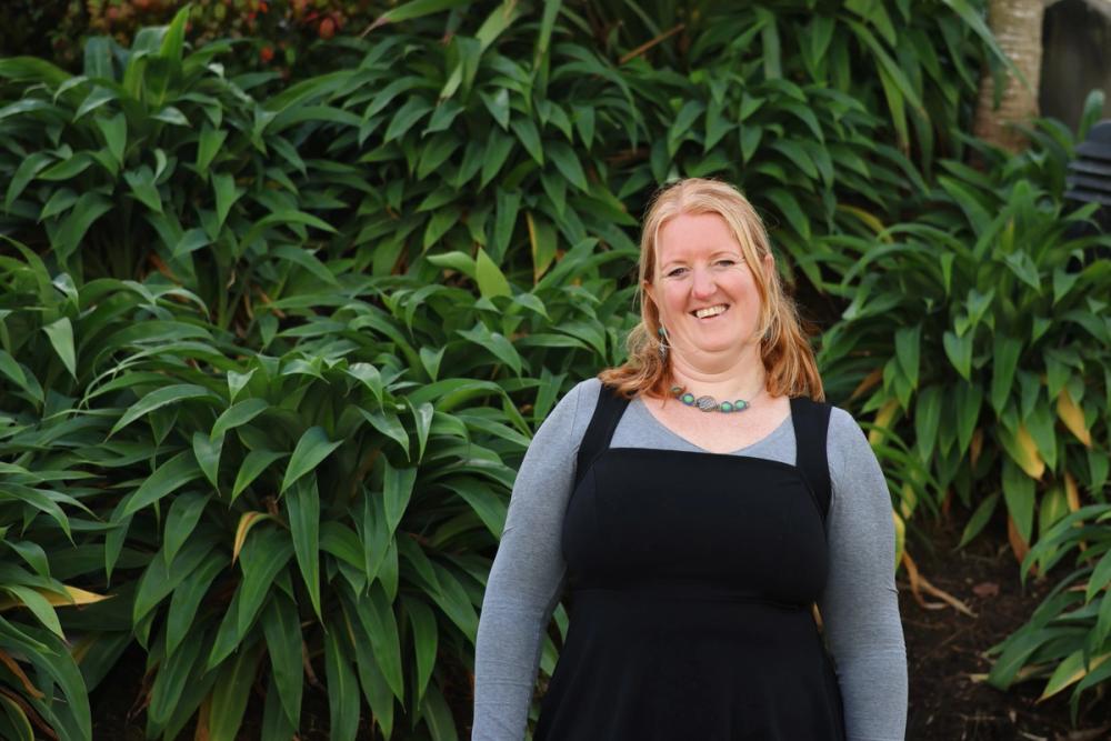 a middle-aged woman standing front of a green leafy bush smiling at the camera