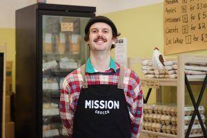 a man wearing an apron is standing in front of a refrigerator in a grocery store .