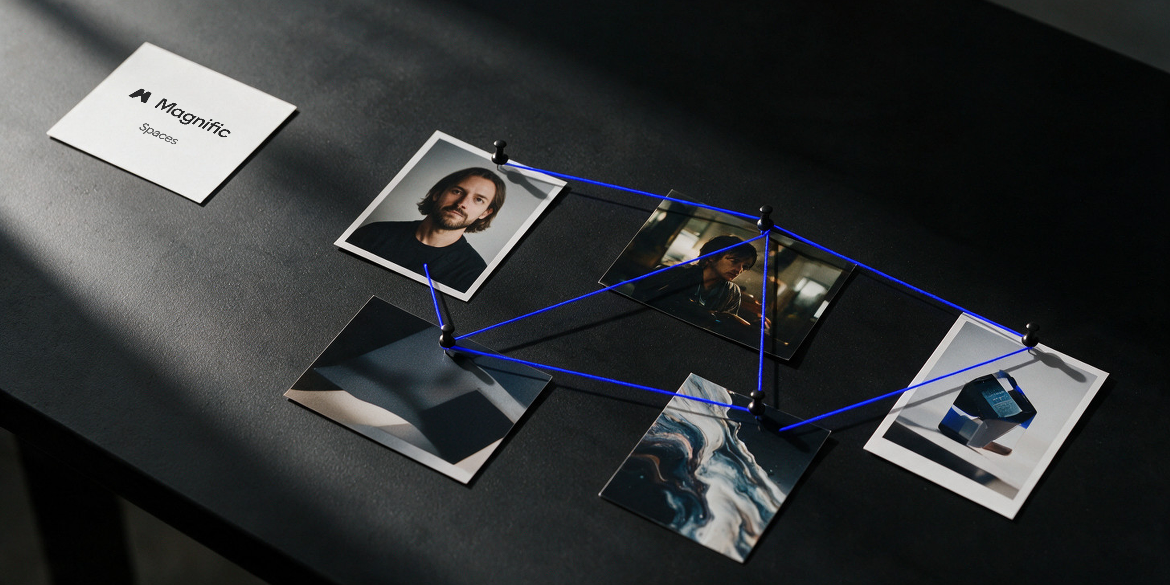 Overhead view of a black desk with printed photos pinned together by electric blue threads like nodes in a workflow, next to a card with the Magnific Spaces logo