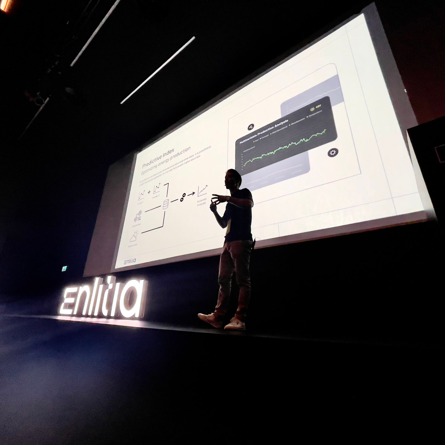 Photograph of one of the team members of Enlitia speeking at the company presentation. In the background, you can see a slide of a presentation, next to the person a neon light in form of the Enlitia logo.