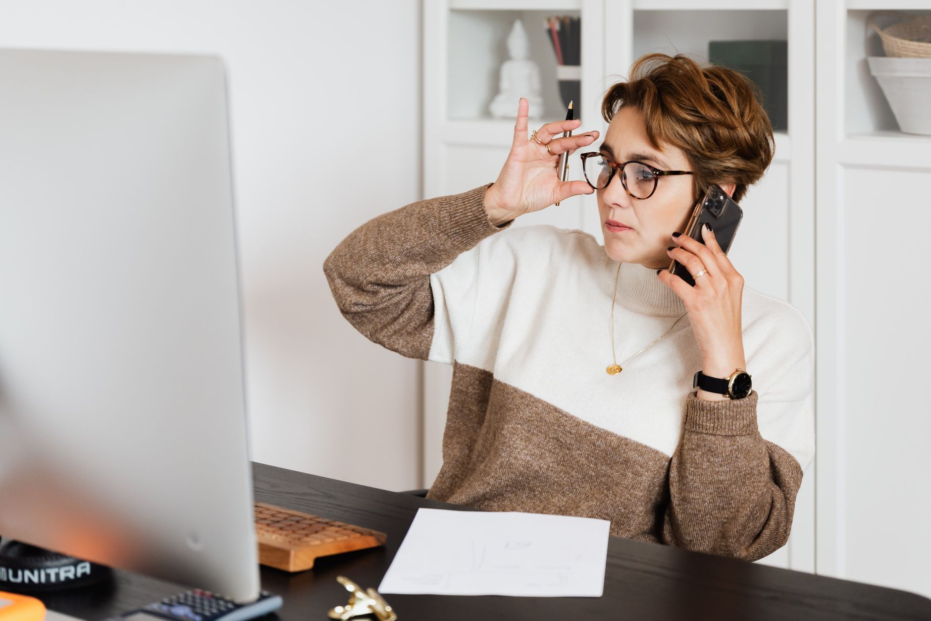 A focused office worker on the phone at her desk, looking at a printed invoice while checking her computer screen, representing the moment a small business confirms a supplier's payment details before processing a payment