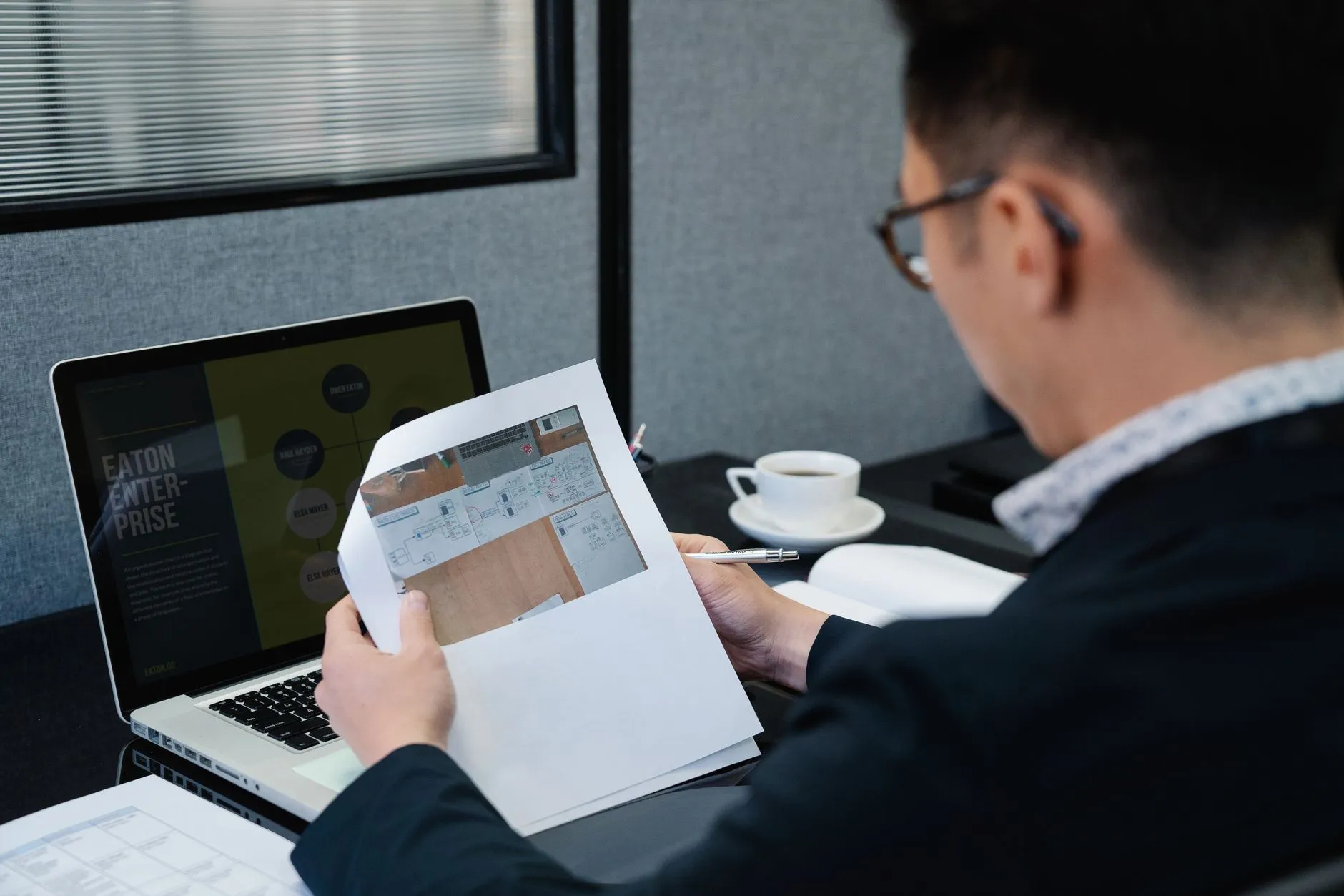 A man at an office desk working through printed documents beside a laptop — the kind of line-by-line review a bank's third-party risk analyst gives a partner's penetration test report before accepting it
