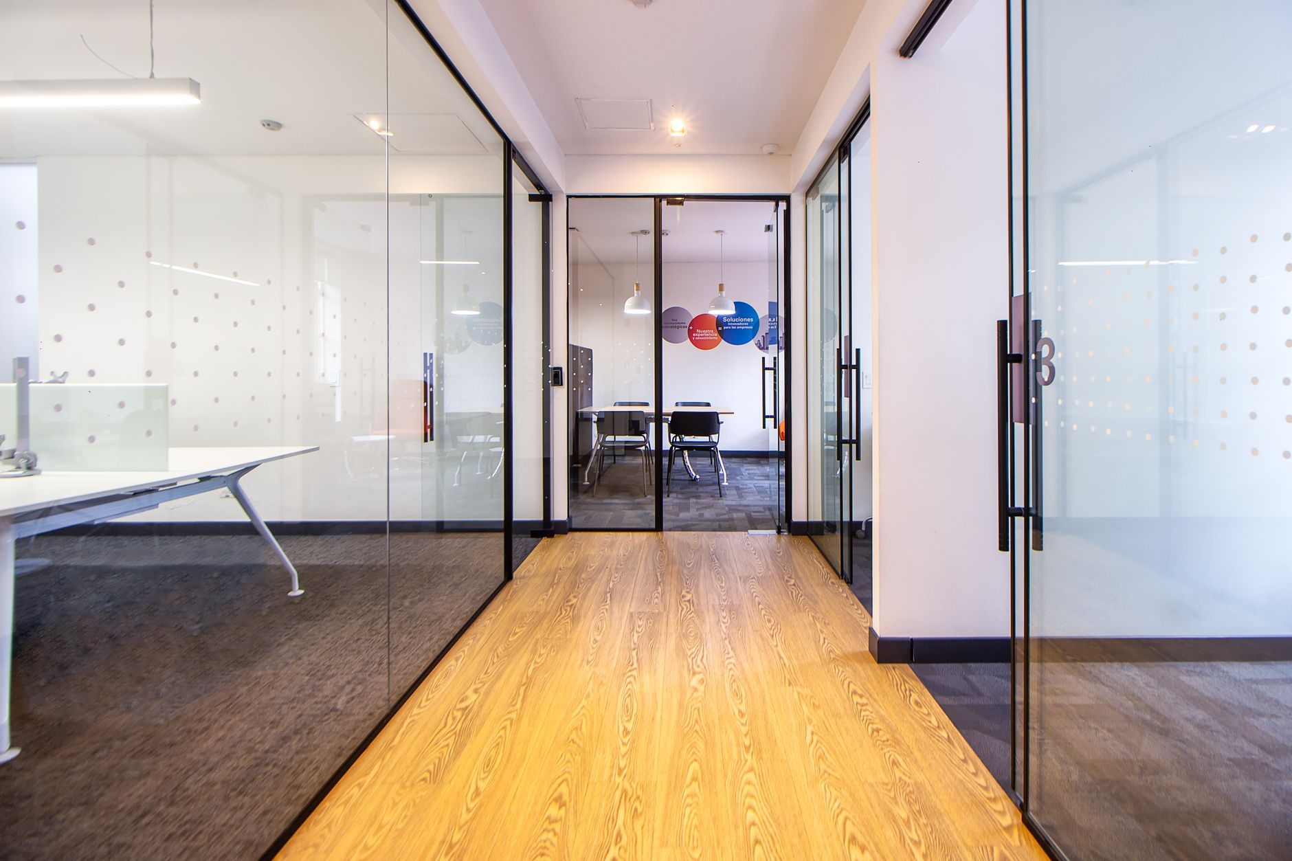A bright office corridor lined with glass-walled meeting rooms — the reflective, partitioned environment on a professional-services floor where two engineers surveying the same space can reach opposite conclusions