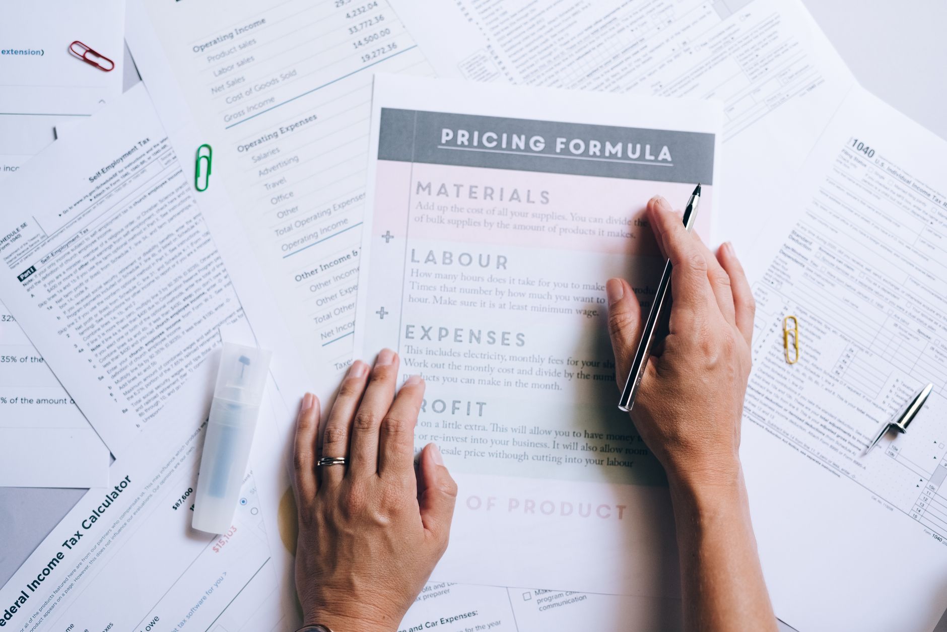 Hands laying out costing documents and a worked pricing formula across an office desk