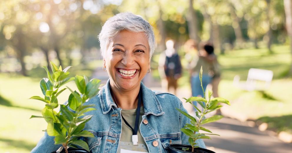 Photo of a woman of colour holding a couple of plants and smiling. She has short grey hair and is standing in a park on a sunny day.