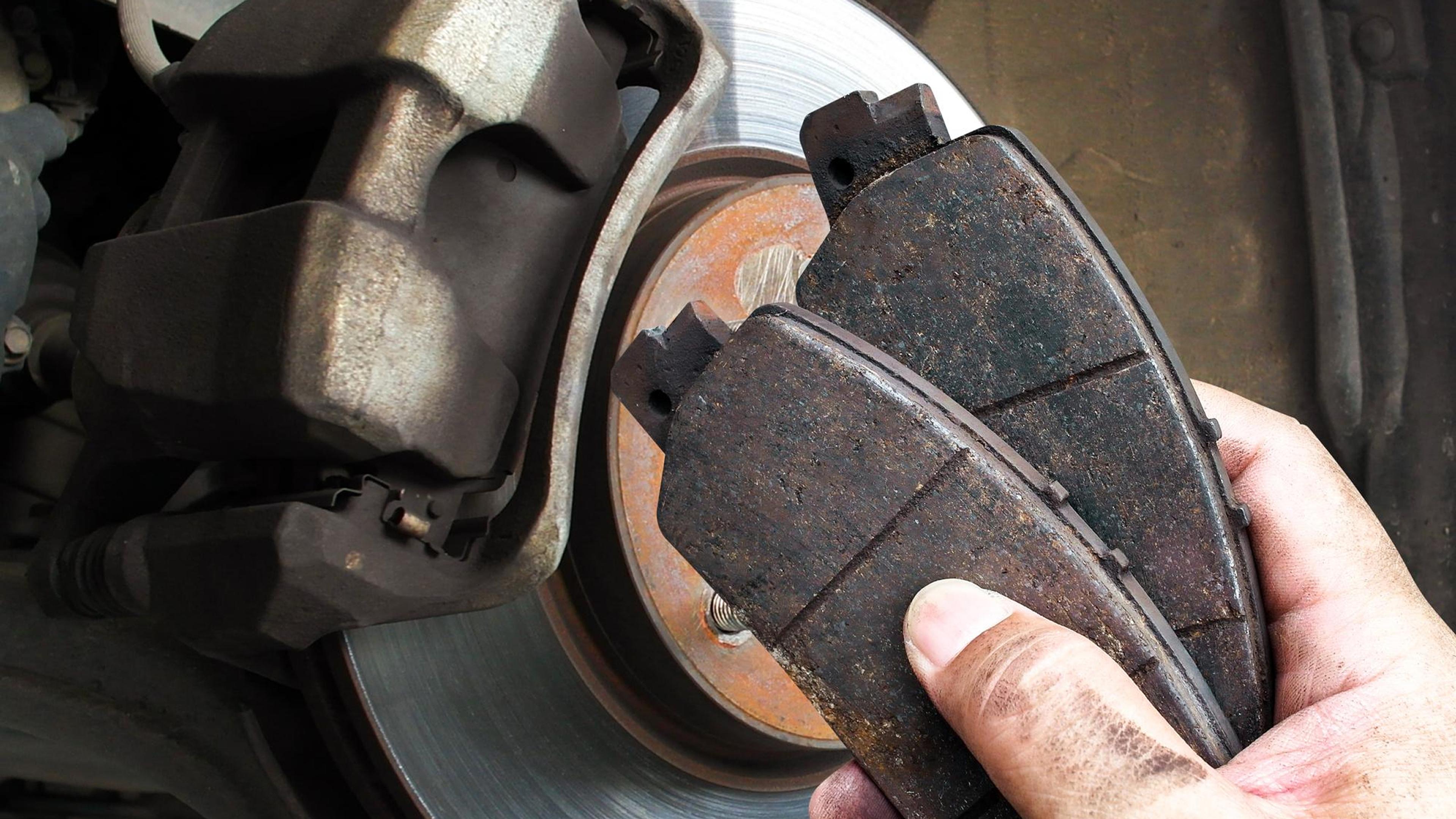 Hand holding worn brake pads in front of a brake disc on a vehicle