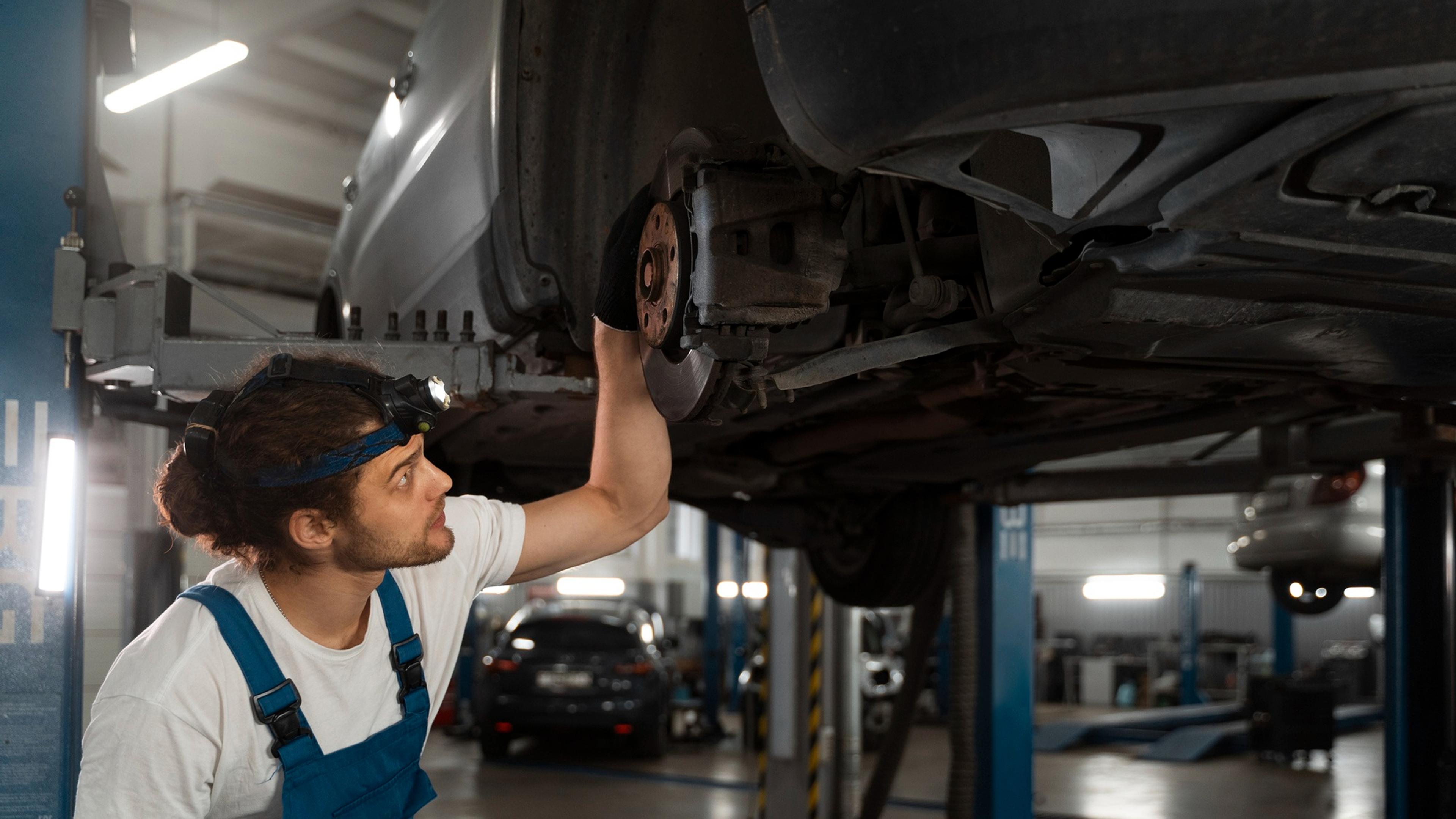 A car mechanic inspects the underside of a raised car in a modern workshop equipped with lifting platforms.