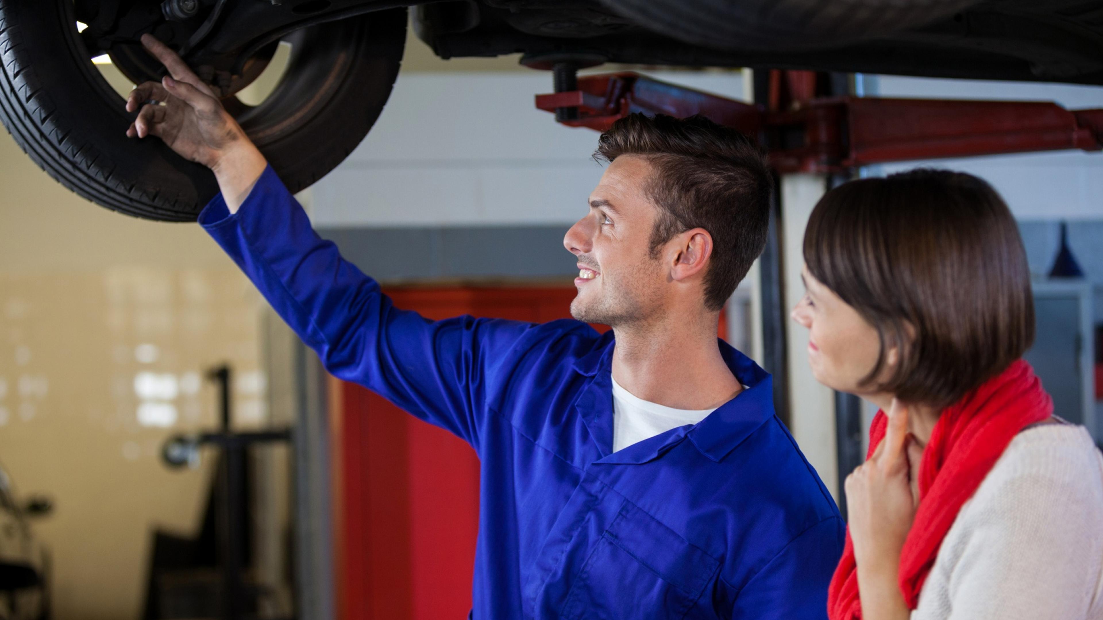 Automotive mechatronics technician explains the condition of the tires and suspension to a customer while the vehicle is raised on a lift.
