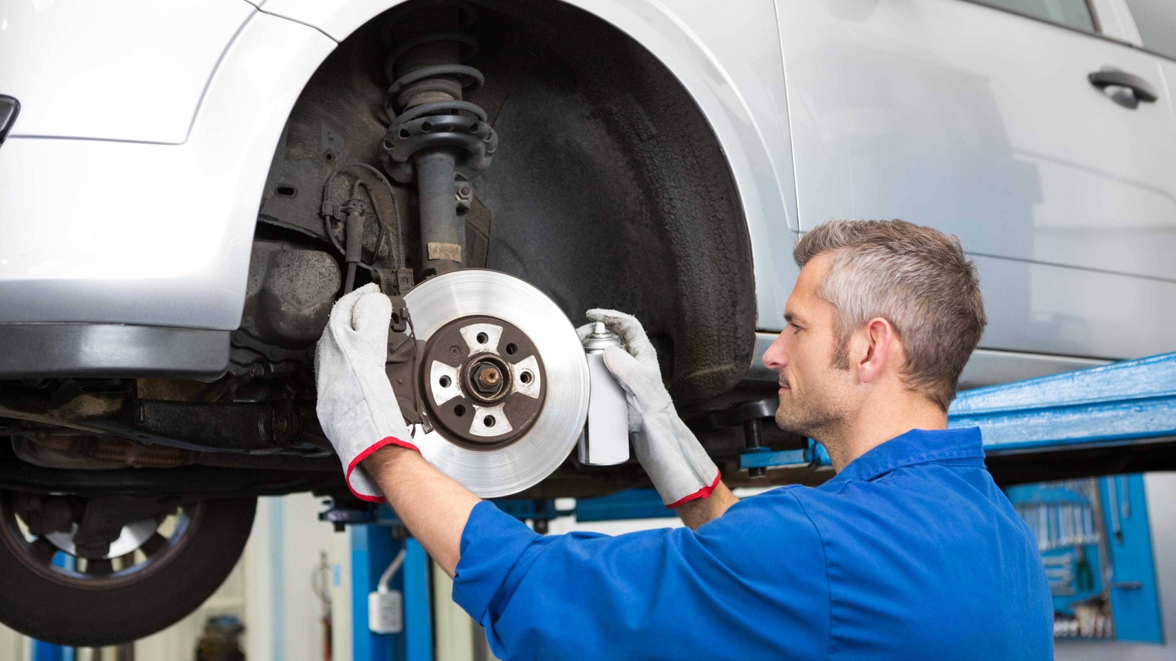 Kfz-Mechaniker reinigt eine Bremsscheibe mit Spray an einem angehobenen Auto in einer modernen Werkstatt.