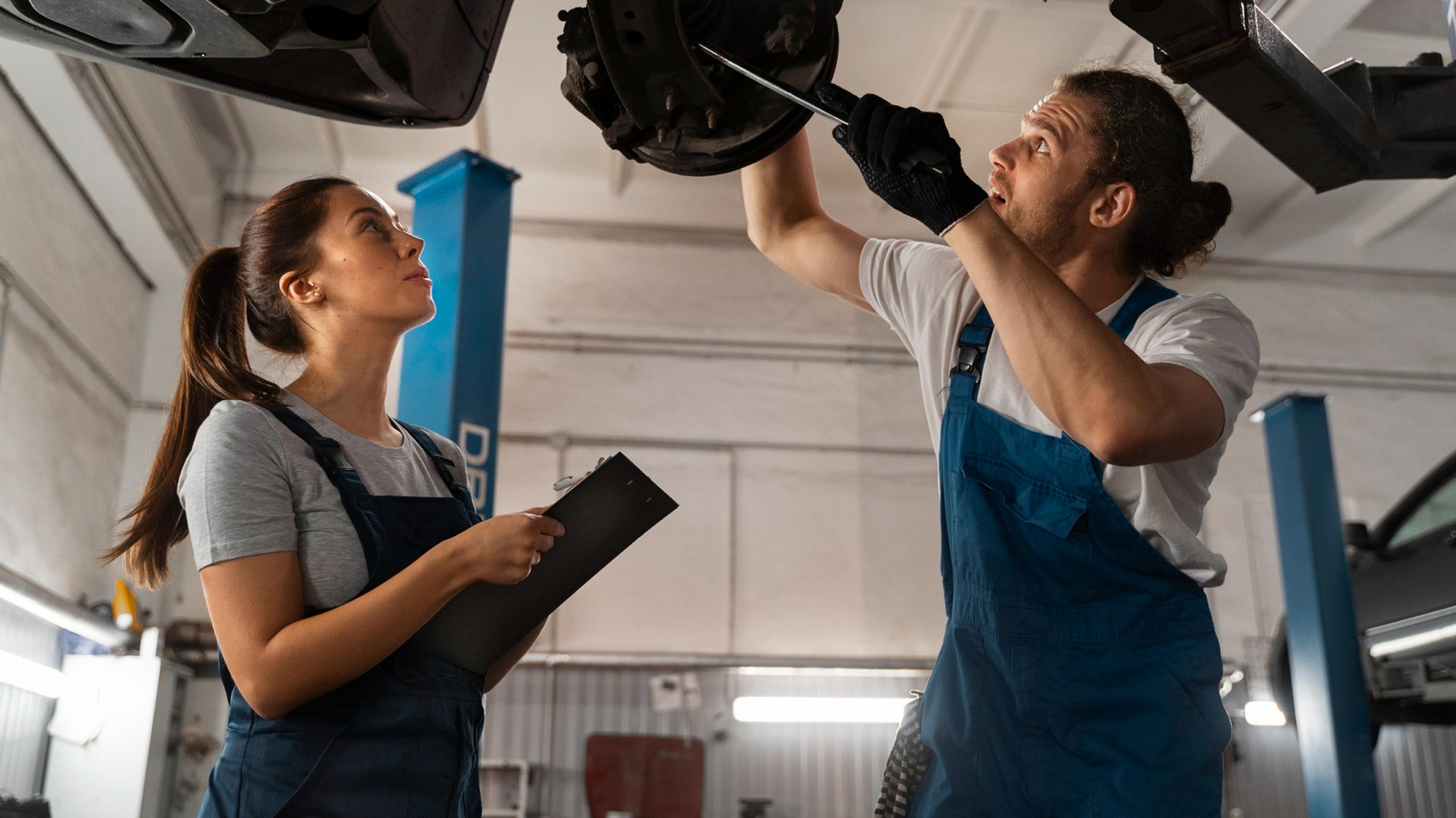 A female mechanic with a clipboard and a male mechanic jointly check the brake system of a raised car in a workshop.