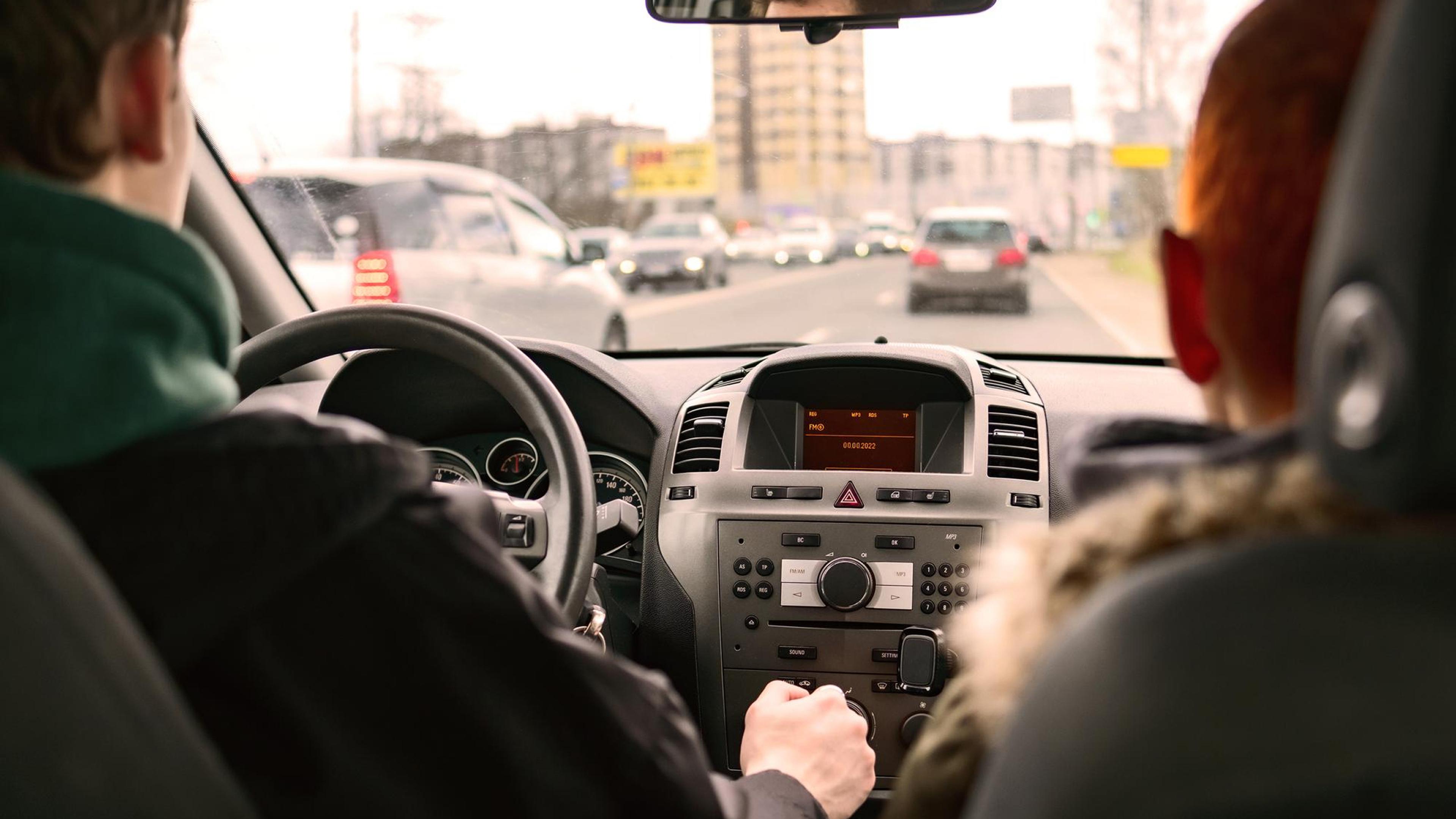 Blick aus dem Rücksitz auf zwei Personen im Auto, während der Fahrer den Verkehr in einer Stadtstraße beobachtet.