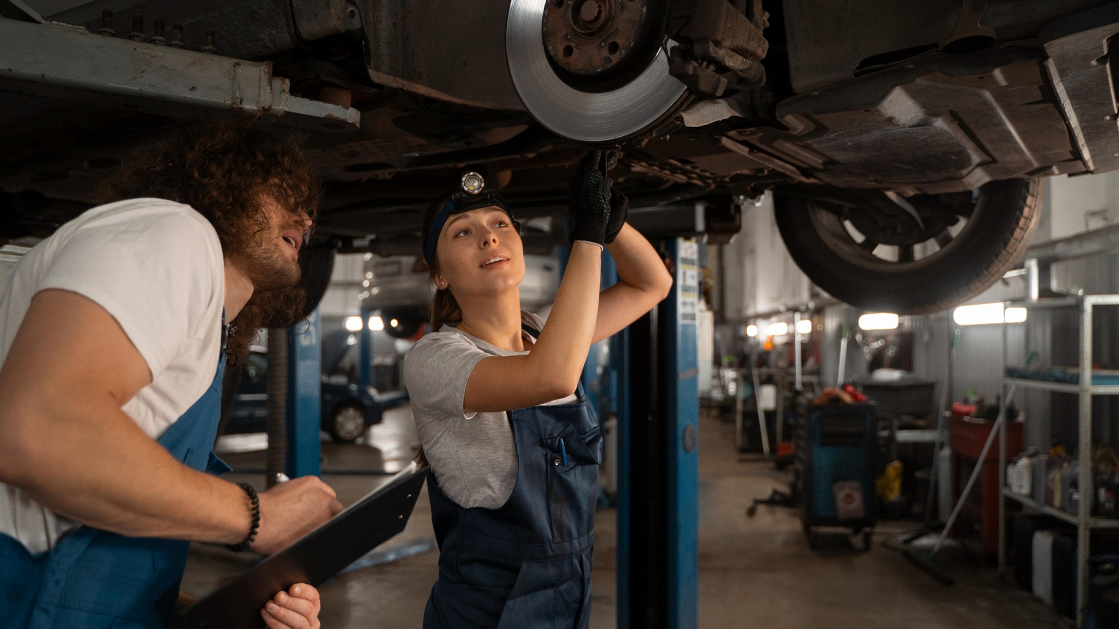 Zwei Kfz-Mechaniker arbeiten unter einem angehobenen Fahrzeug und kontrollieren die Bremsanlage in einer Autowerkstatt.