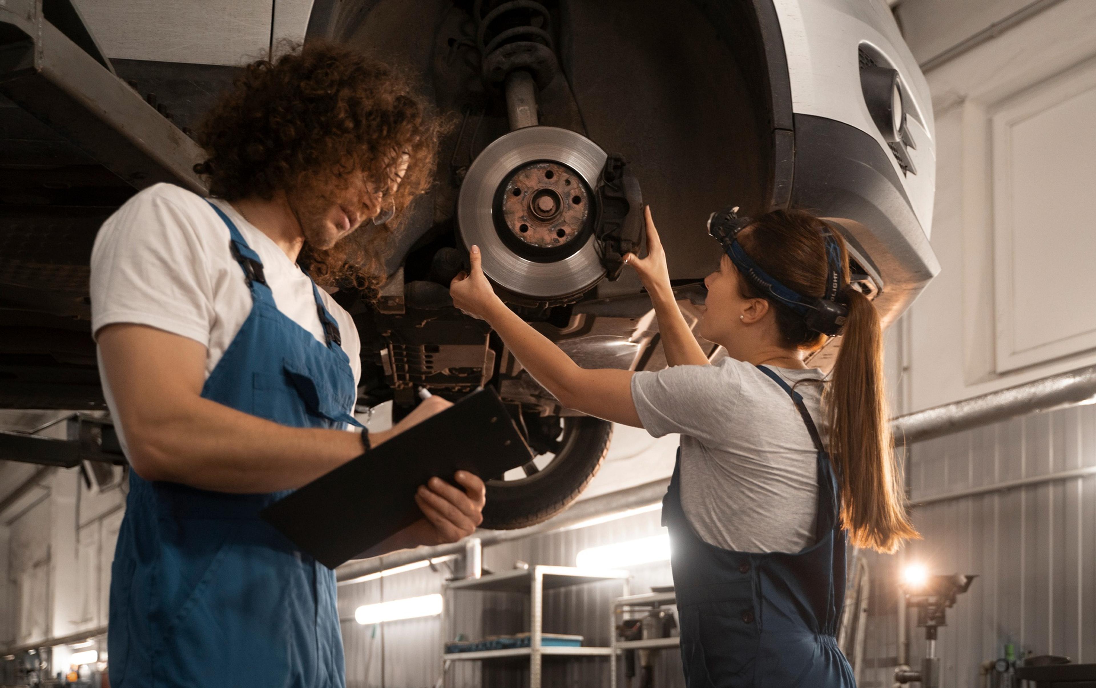 Mechanikerin montiert eine Bremsscheibe an einem angehobenen Auto, während ein Kollege mit Klemmbrett die Arbeit überprüft.