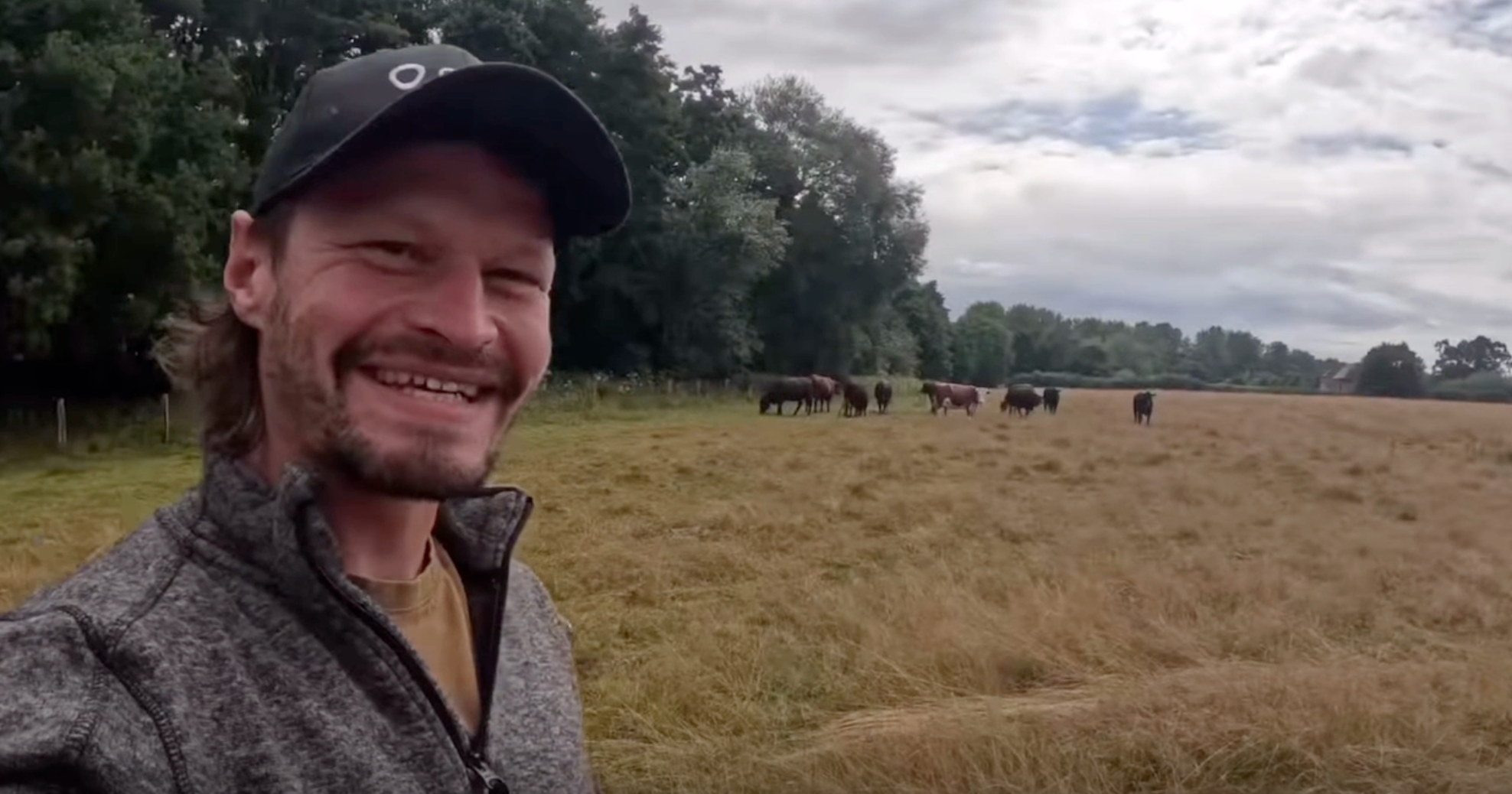 Chris smiling with his cows behind him wearing their Monil Virtual Fencing Cattle Collars.