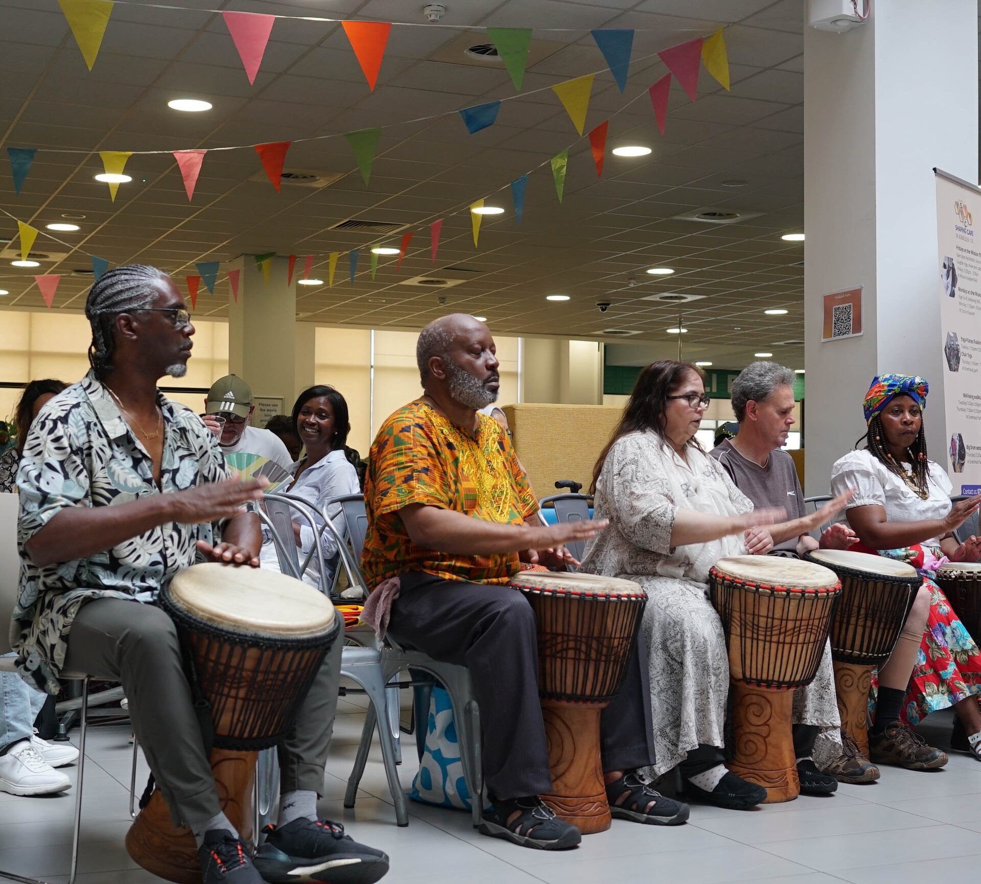 Multi-generational community drumming session with djembe drums beneath colourful bunting.