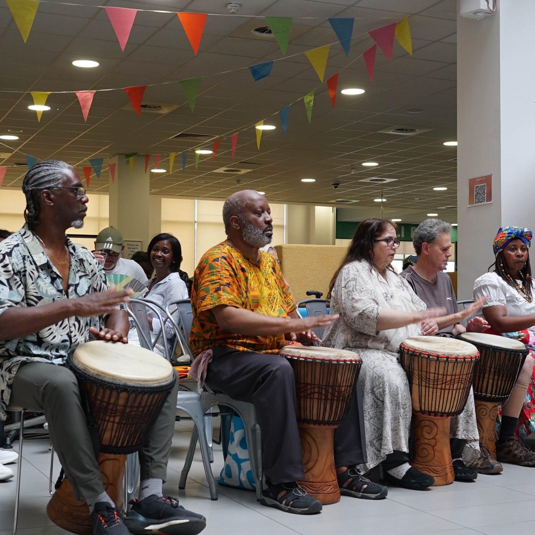Multi-generational community drumming session with djembe drums beneath colourful bunting.