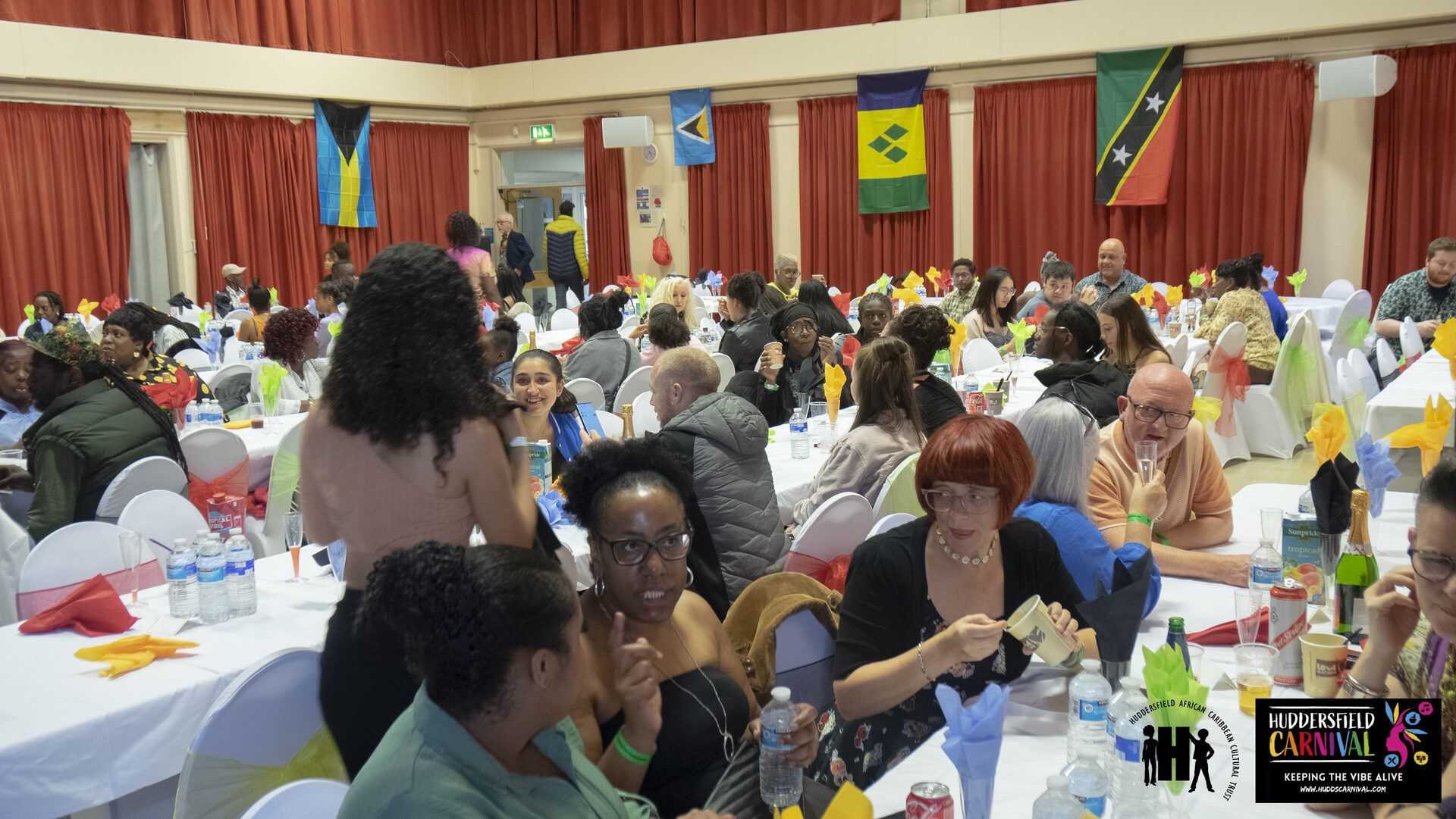 A community dinner hall with Caribbean flags hung on the wall and tables of guests.