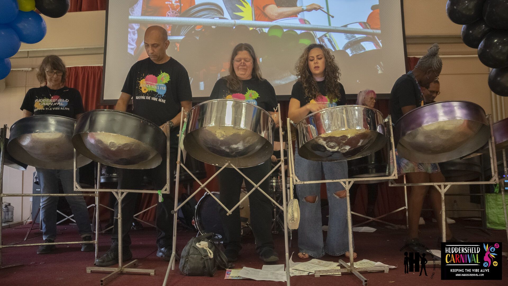Five members of the Legacy Steel Band performing on stage in matching black HACCT t-shirts, with steel pans on stands.