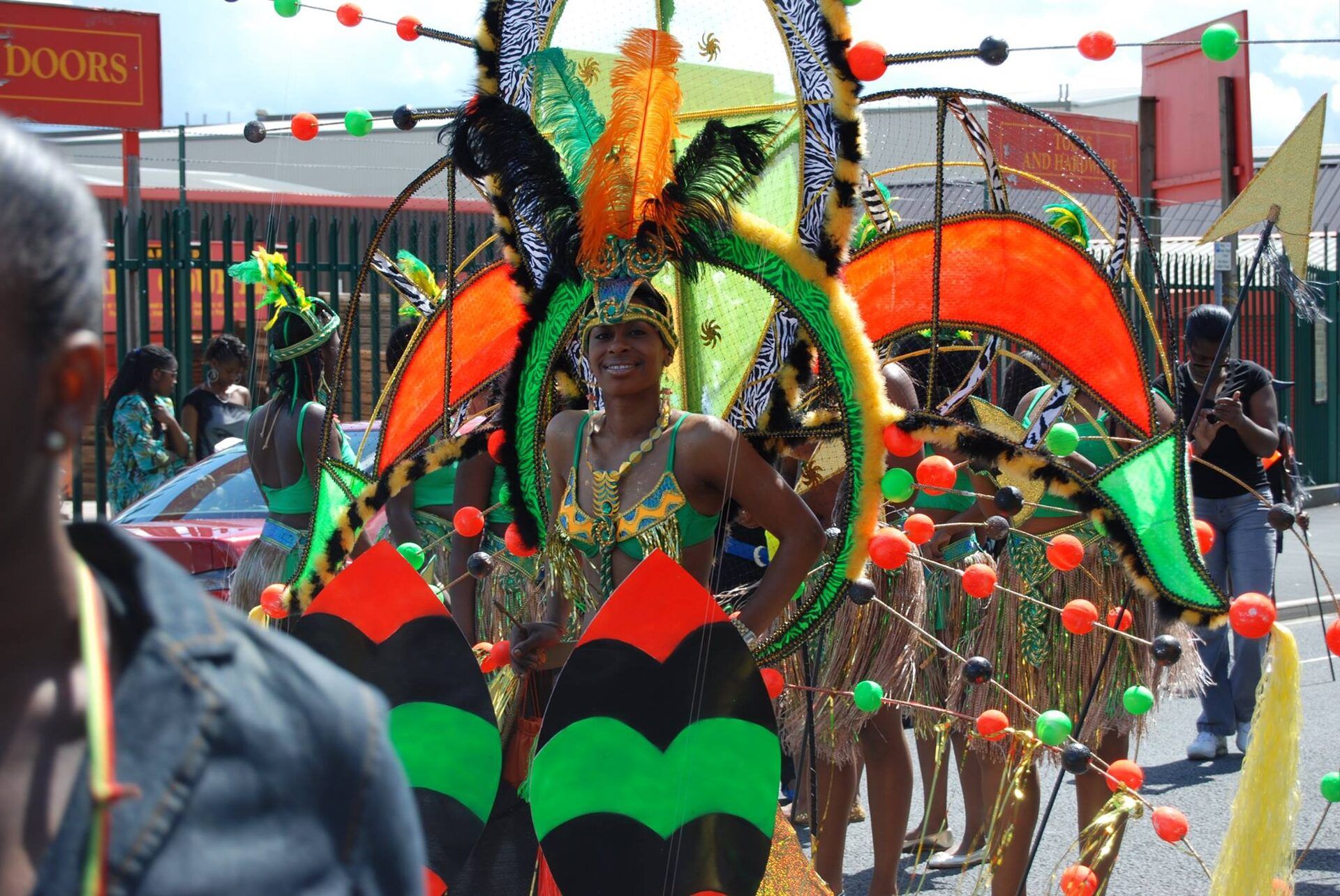 A Carnival performer in full costume — a sunburst headdress in green, orange and gold, with feathers and beadwork.
