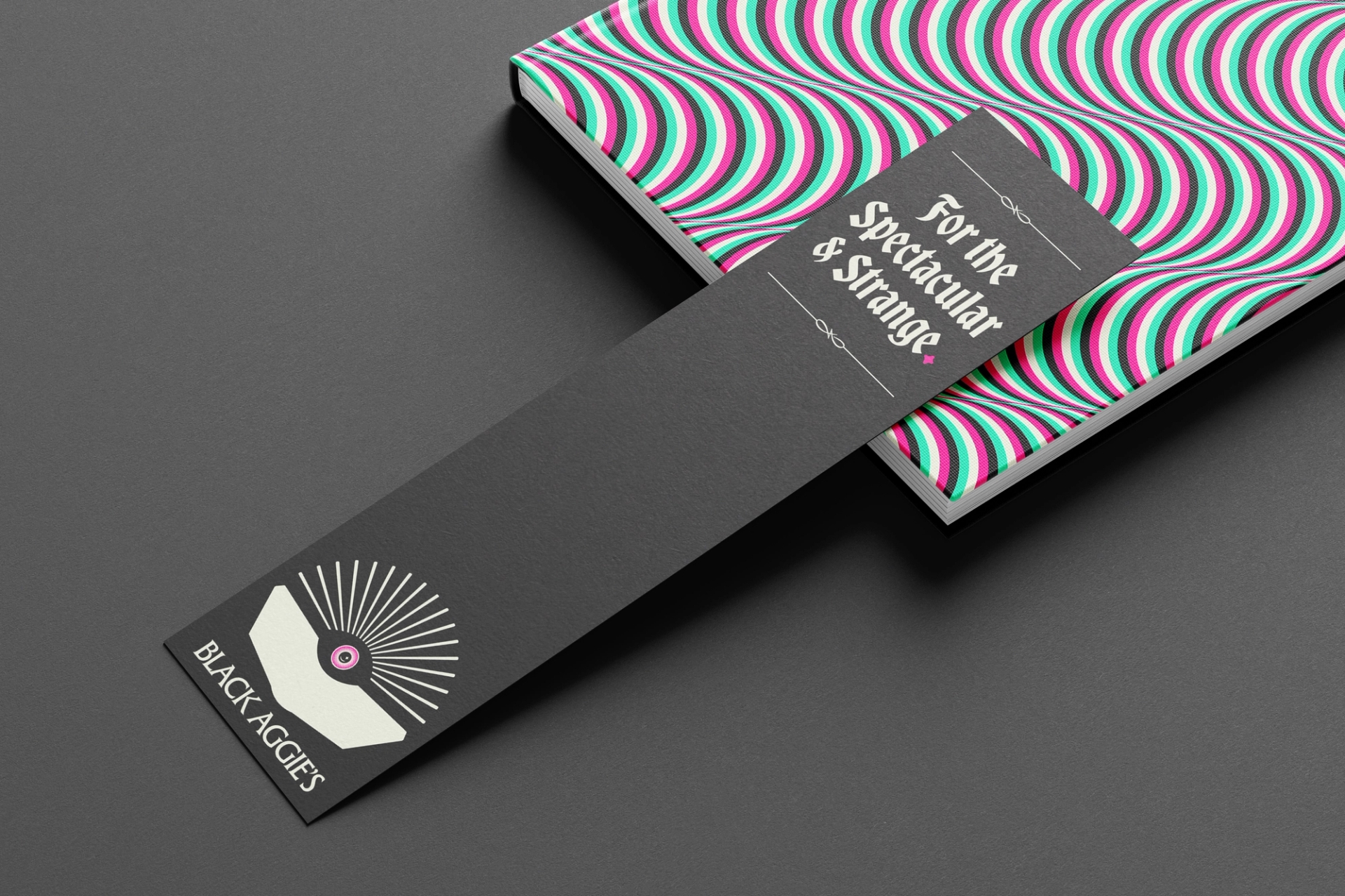 Bookmark with Black Aggie's logo leans on a closed book. The bookmark says "For the Spectacular & Strange"