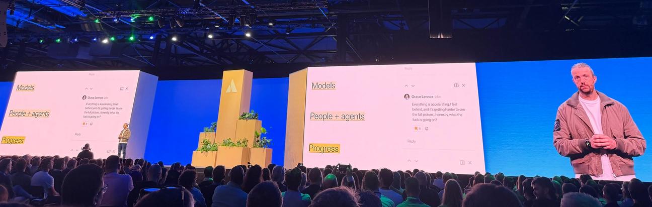 A photo of the screen at the keynote session showing three large titles 'models', 'people and agents', and 'progress'. Alongside the screen stands Mike Cannon-Brookes