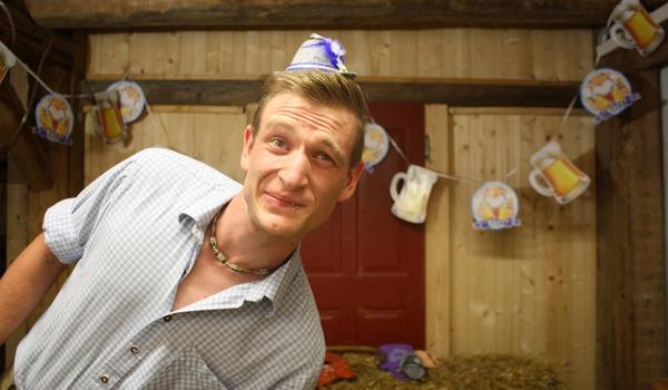 Mein junges Ich lehne mich mit einem verschmitzten Lächeln in der Fotoecke seitlich vor die Kameralinse. Ich trage ein blau-weiß kariertes Hemd und einen winzigen, passenden Deko-Hut Filzhut mit Feder auf dem Kopf. Im Hintergrund ist eine Partygirlande und Heuballen mit weiterer Verkleidung zu erkennen.