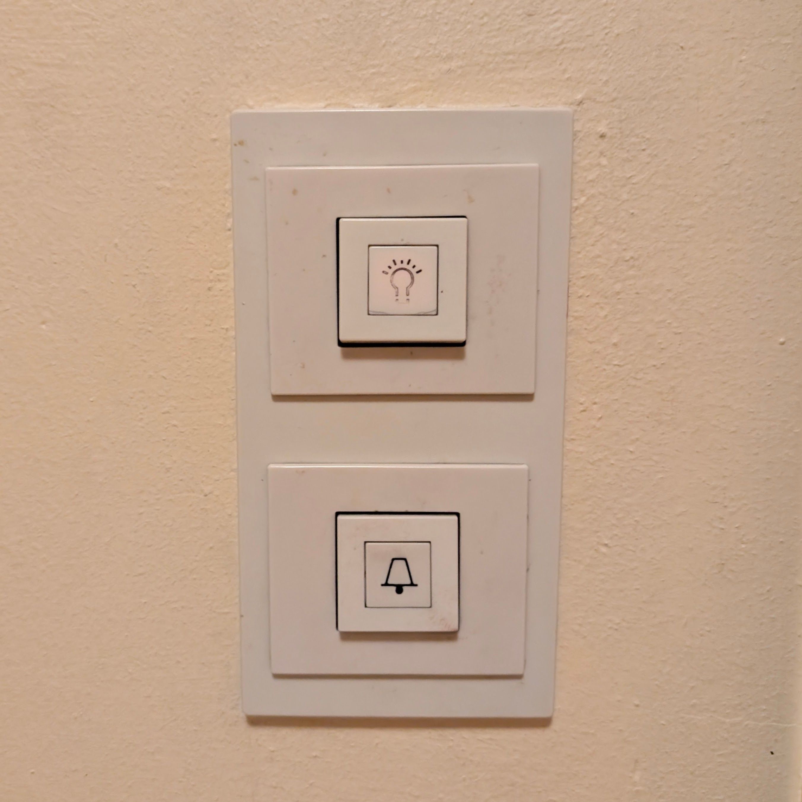 Two white wall switches on a beige wall; the top switch has a light bulb symbol, and the bottom switch has a doorbell symbol.