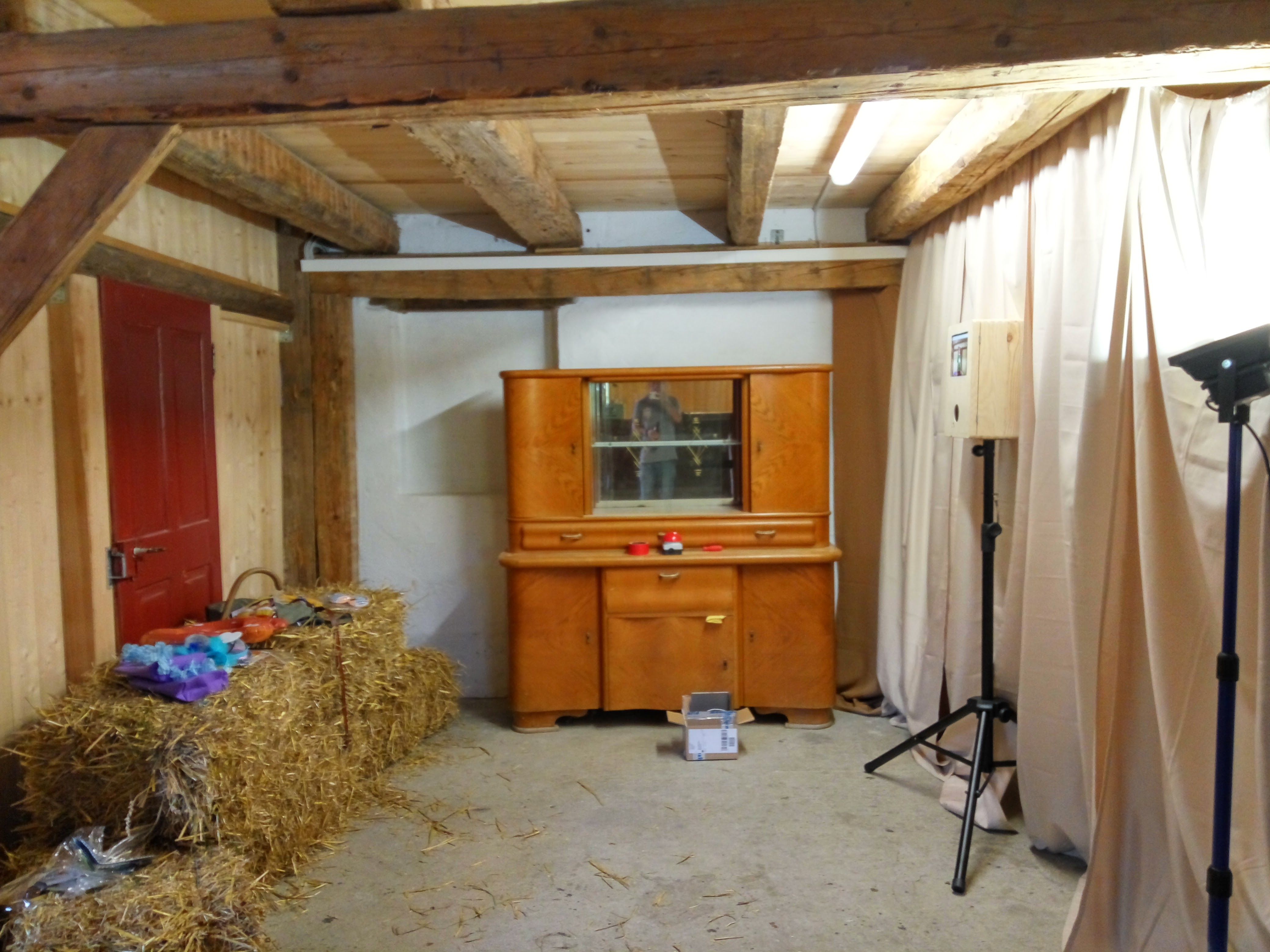 Aufbau einer kompletten Fotoecke in einer rustikalen Holzscheune mit sichtbaren Deckenbalken. Im Zentrum steht ein Vintage-Sideboard aus Holz mit Spiegel, links davon als Hintergrund liegen Heuballen mit einem Korb voller bunter Verkleidungen. Rechts sind die hölzerne Fotobox und ein Scheinwerfer auf Stativen aufgestellt, eingerahmt von einem hellen Vorhang.