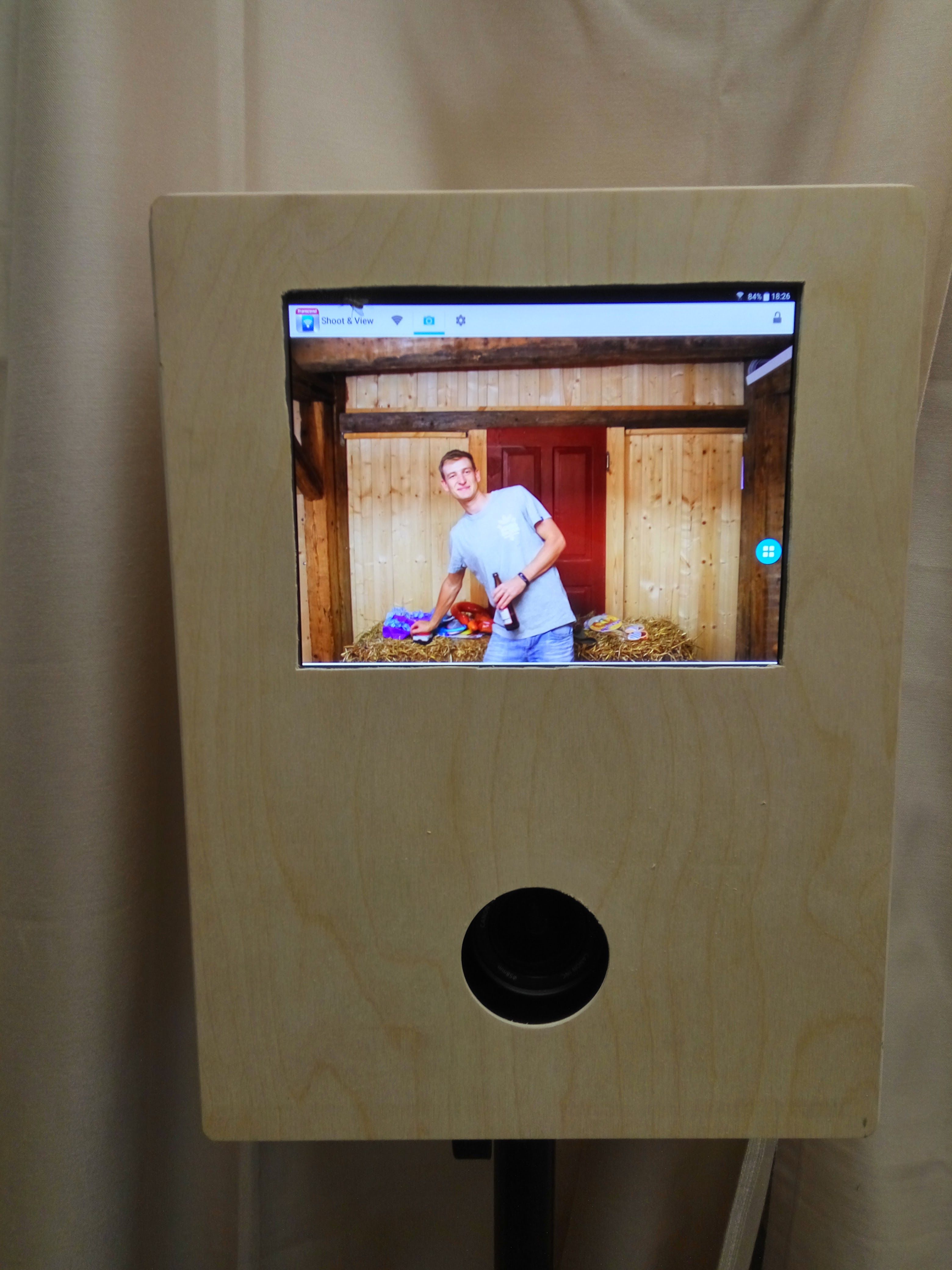 Gehäuse der aufgestellten Fotobox auf einem Stativ von vorne. Das schlichtes Gehäuse aus hellem Holz hat oben eine Aussparung für den Bildschirm, darauf bin ich in einer Scheune, und darunter ein kreisrundes Loch für das Kameraobjektiv.
