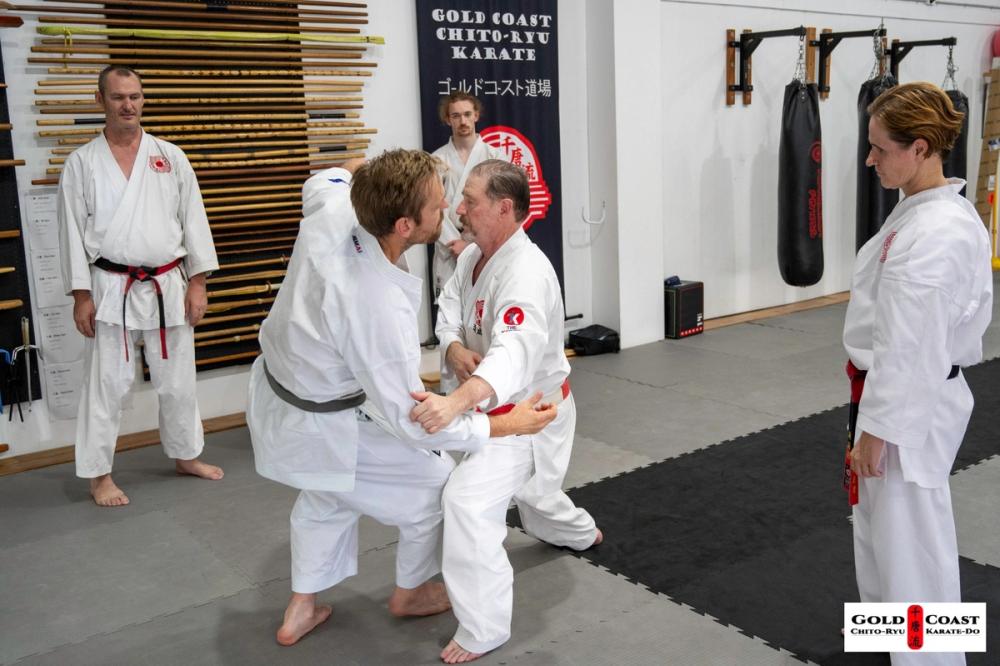 Karate students practicing grappling in the dojo while instructors observe.
