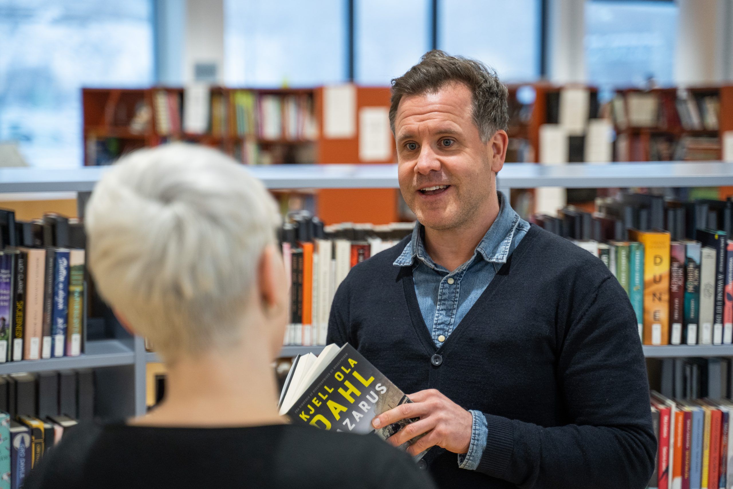 En mann holder en bok og snakker med en kvinne i et bibliotek.